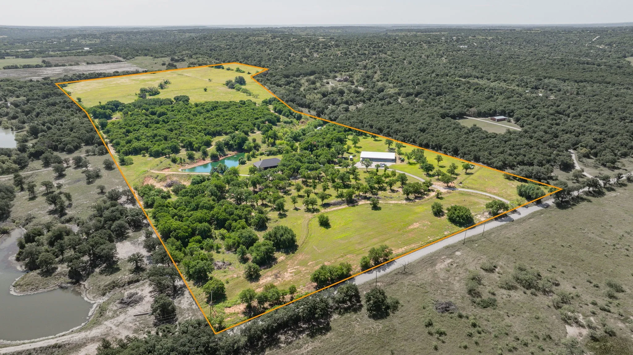 Expansive parcel featuring a large structure, multiple outbuildings, a private pond, abundant mature trees, and open fields, bordered by a paved road