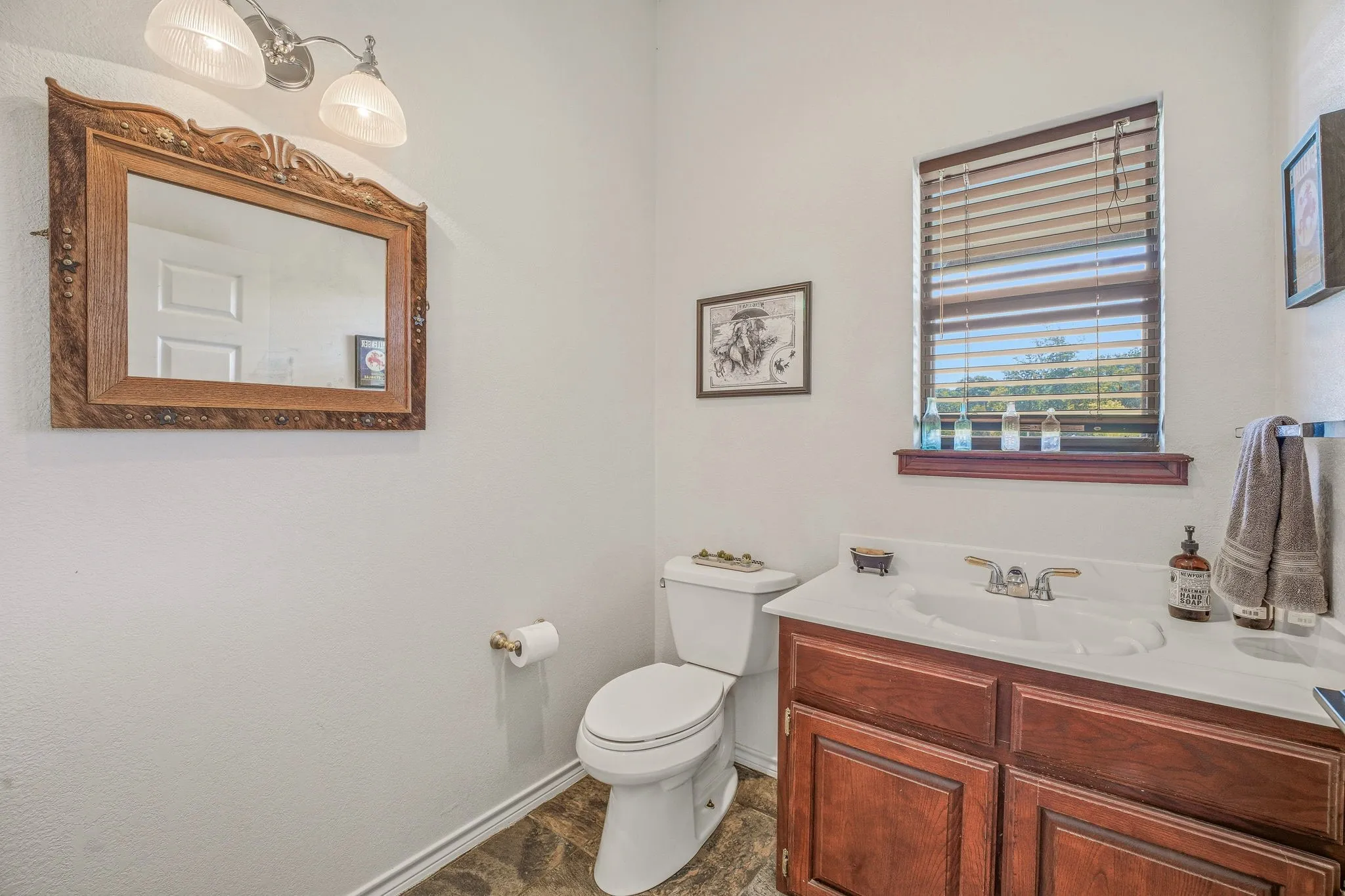 Bathroom featuring a wood-finish vanity with an integrated sink and white countertop, wood-finish flooring, a window with horizontal blinds, and a decorative mirror with an ornate frame