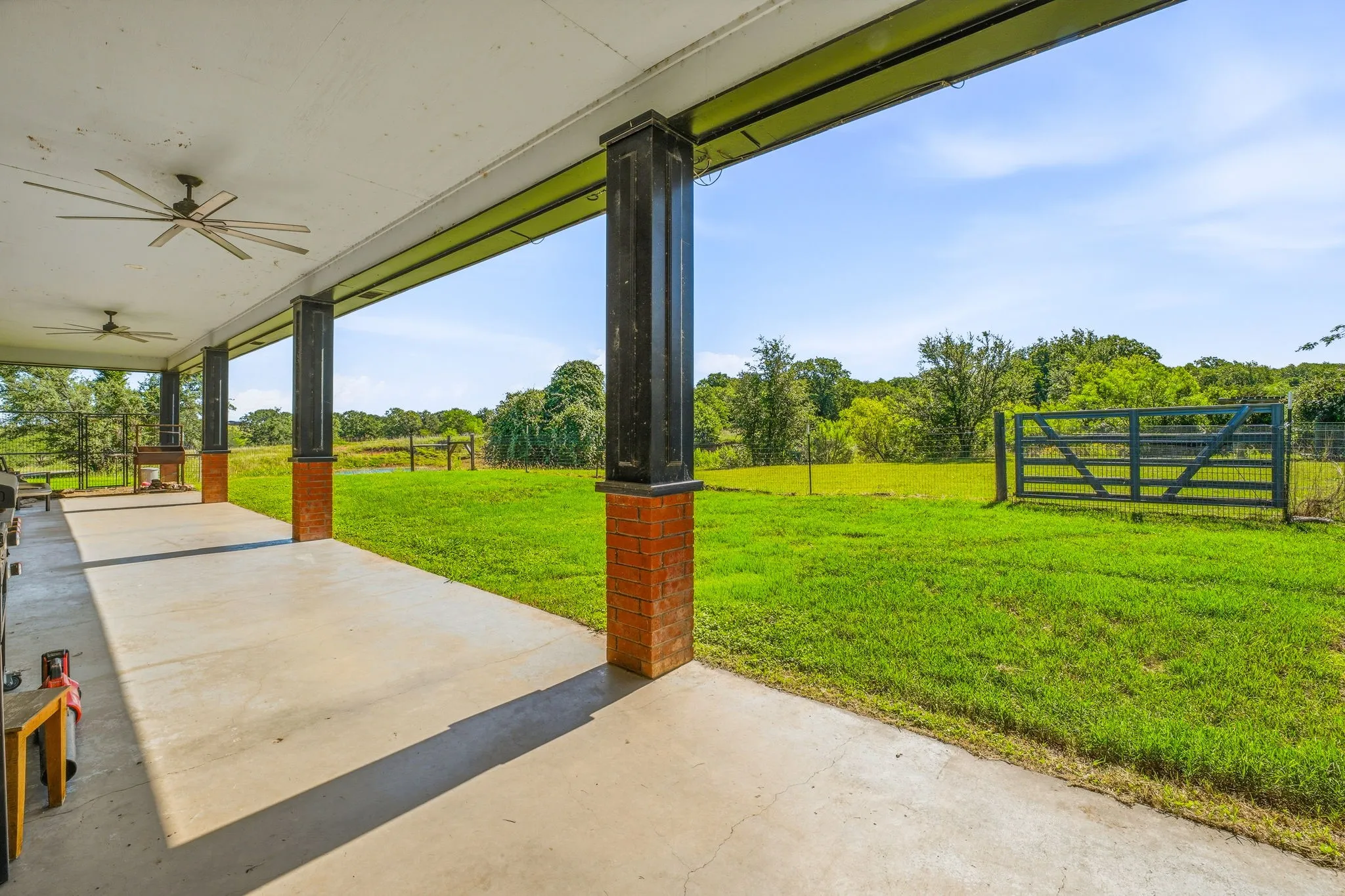 Expansive covered patio featuring a concrete surface, multiple ceiling fans, brick-clad columns, and an open-air built-in grill station, overlooking a sizable grassy area with perimeter fencing and a wooden gate