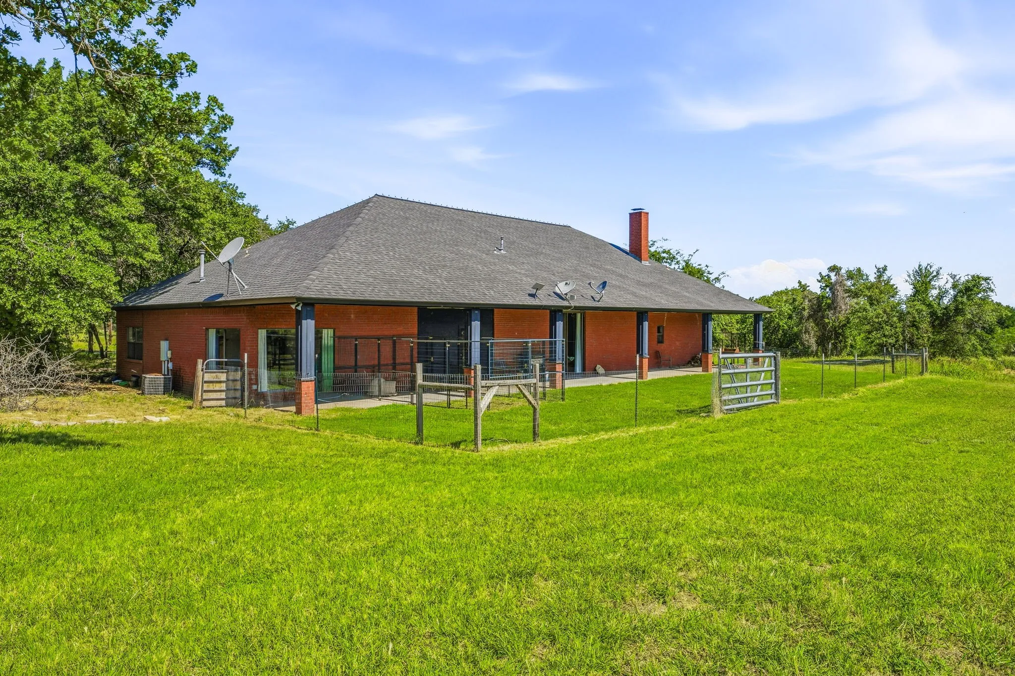 Expansive green lawn surrounds a red-hued exterior home featuring a dark shingle roof, a red brick chimney, and a covered back patio with metal railings