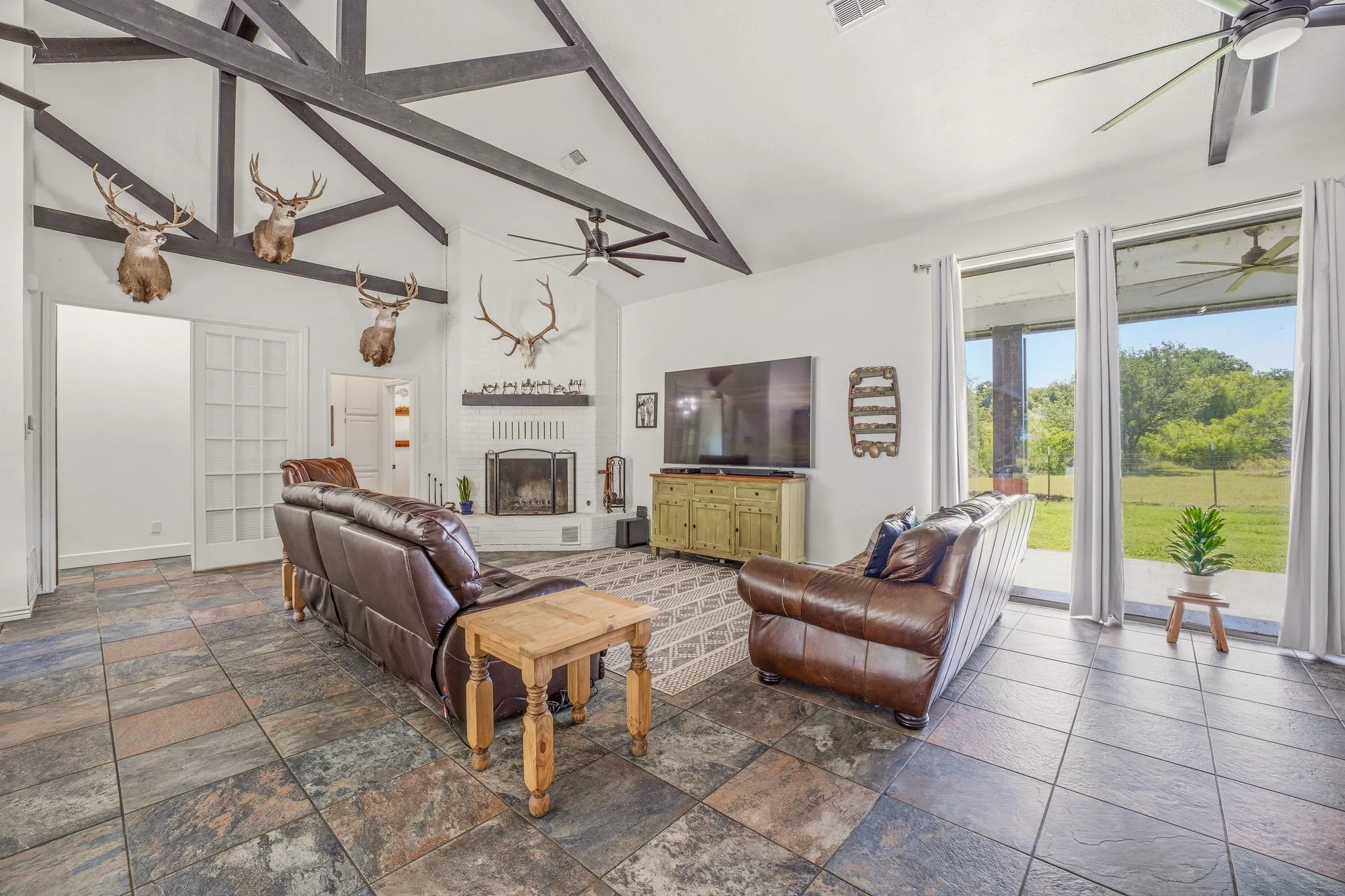 Spacious living area featuring a vaulted ceiling with dark wood beams, a white brick fireplace, and large sliding glass doors opening to the exterior