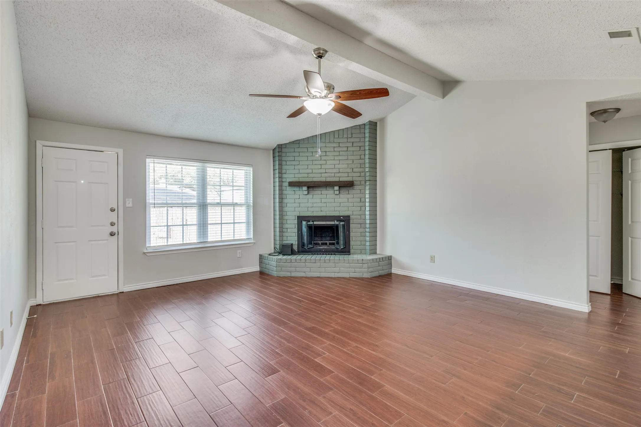 Spacious room featuring vaulted ceilings with exposed beams, wood-finish flooring, and a corner fireplace with a brick surround and wood mantel