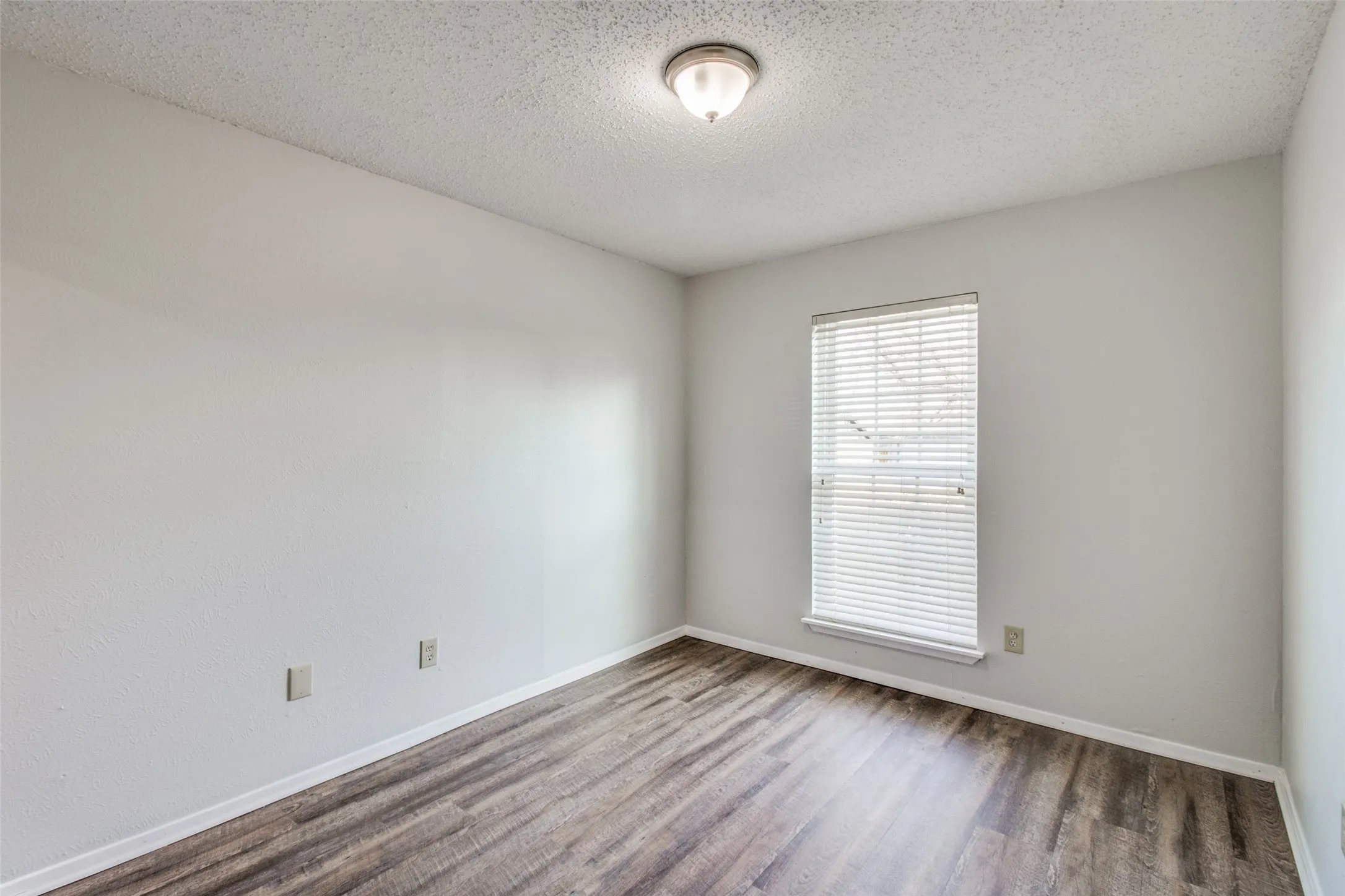 Spacious room featuring wood-finish flooring, light gray walls, a window with horizontal blinds, and a flush-mount ceiling light