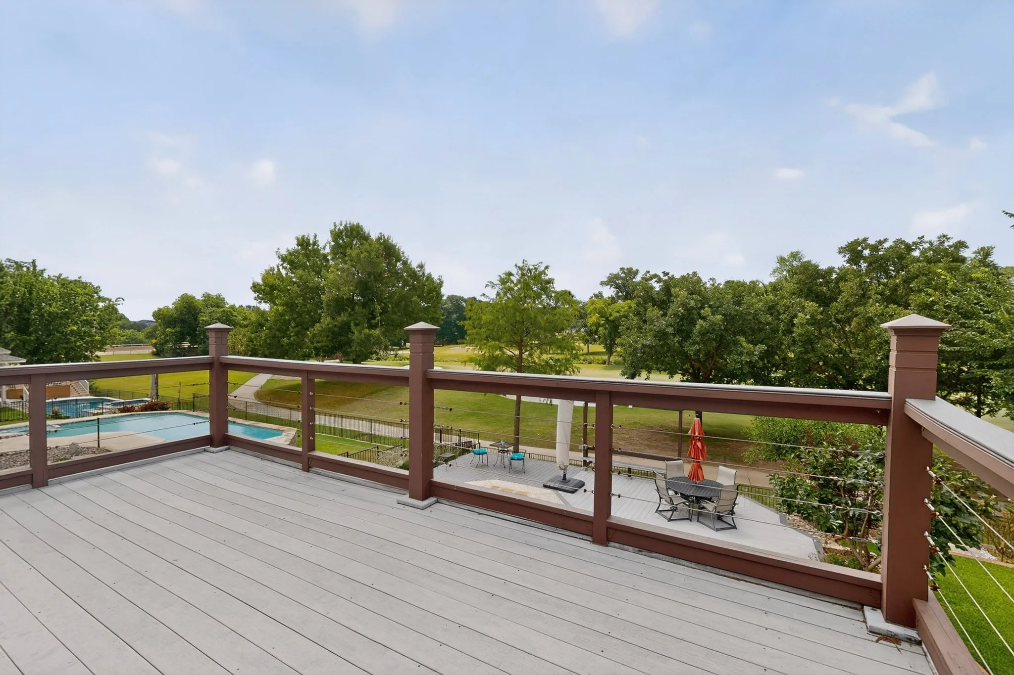 Elevated wood-finish deck with cable railing overlooking a landscaped backyard featuring a swimming pool, concrete patio, and mature trees