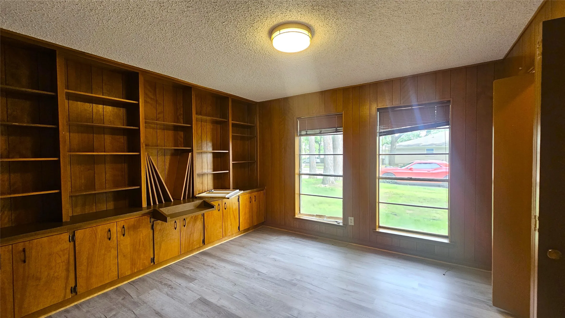 Bedroom 5 with gray luxury vinyl plank floors, windows facing front yard, wall of bookshelves with stained wood and storage. Would be great study or teaching room as well!