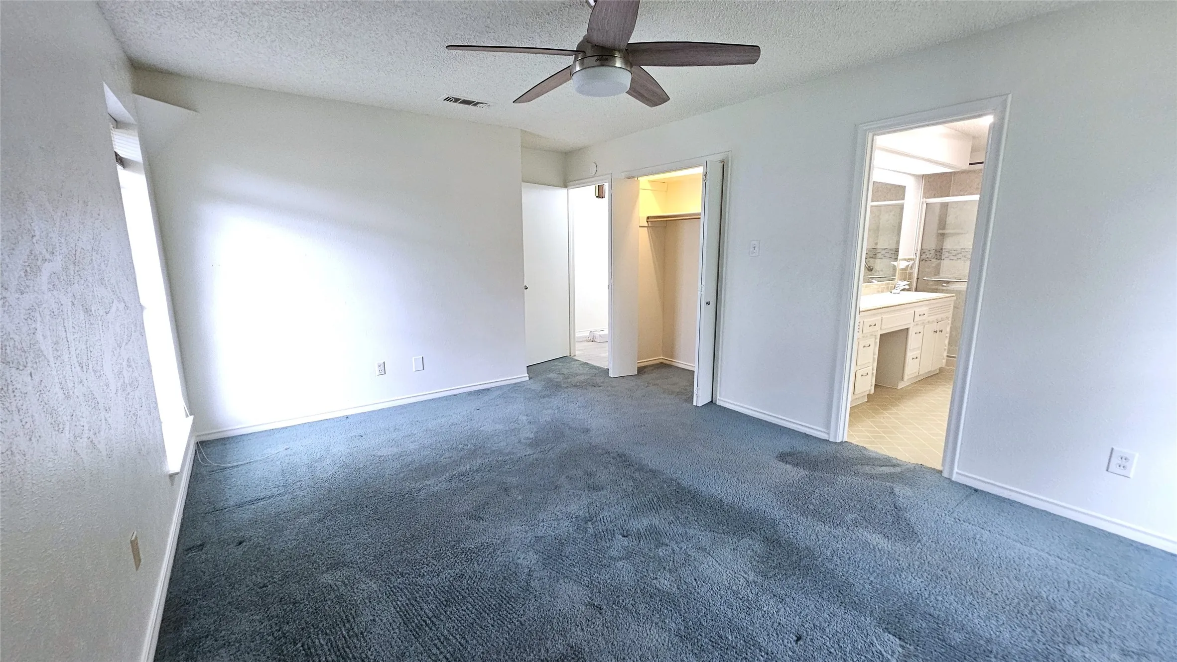 Primary bedroom with ensuite full bath, blue carpet, healthy amount of natural light form windows facing side and back yards, a textured ceiling, ceiling fan, and hidden accent corner lighting.