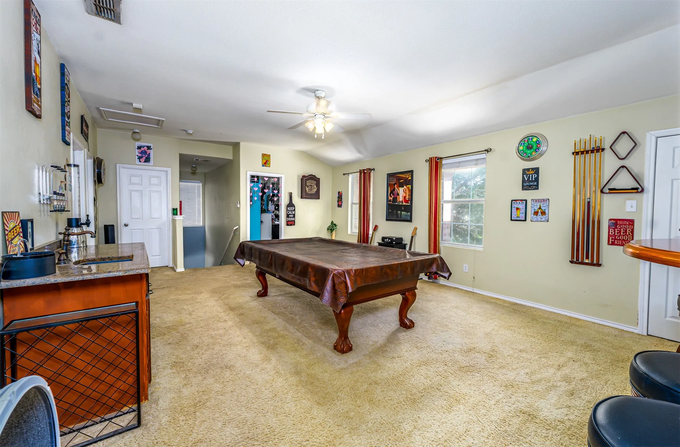 Spacious recreation room featuring a pool table, textured carpeting, two windows with red drapes, and a ceiling fan