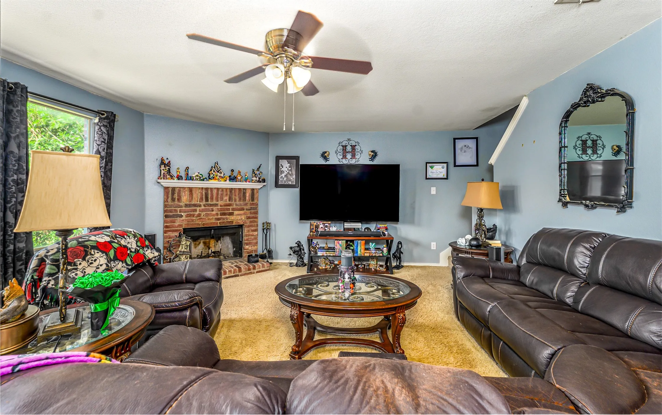 Living area featuring a brick fireplace with a raised hearth, a ceiling fan with integrated lighting, and a large window with dark drapery