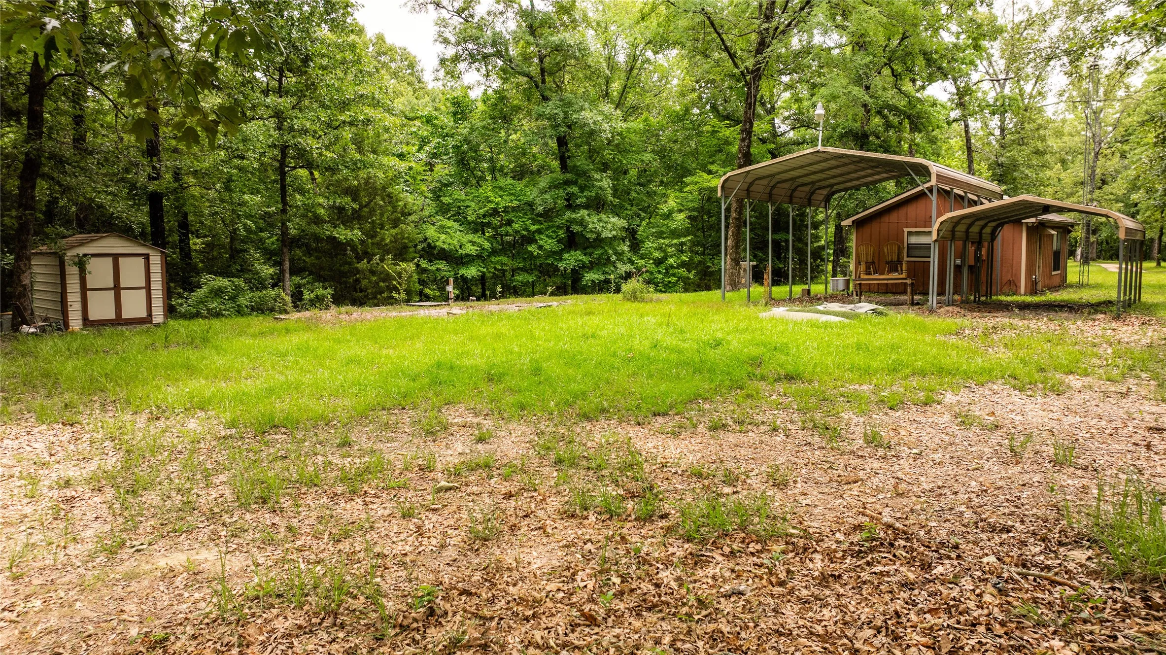 Expansive outdoor space featuring a detached storage shed, multiple metal-frame carports, and a wooded perimeter