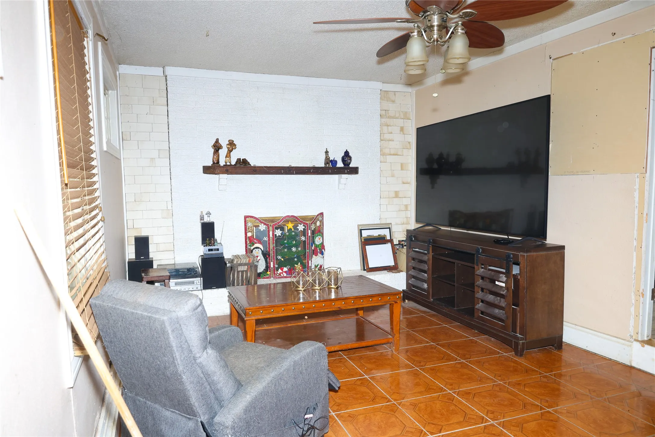 Living area featuring light-toned tile flooring, a painted brick fireplace with a wood mantel, and a ceiling fan with integrated lighting