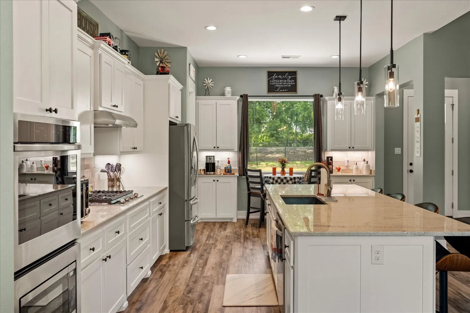 Spacious kitchen featuring white cabinetry, wood-finish flooring, a large center island with an undermount sink, stainless steel appliances, and pendant lighting