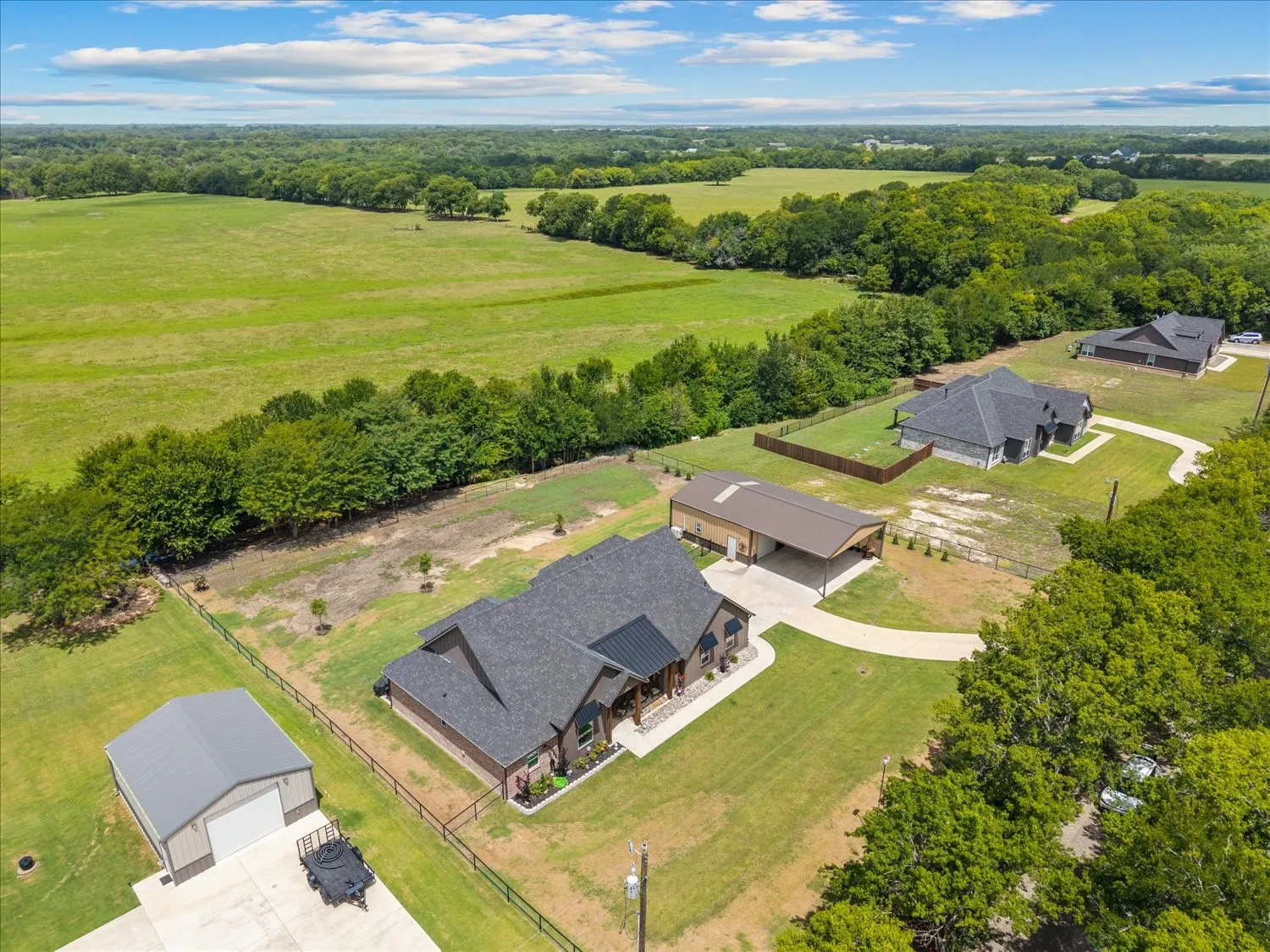 Modern farmhouse-style residence with dark gray roofing and a wraparound porch, featuring a separate metal outbuilding with a roll-up door, a detached carport, a concrete driveway, and extensive green landscape