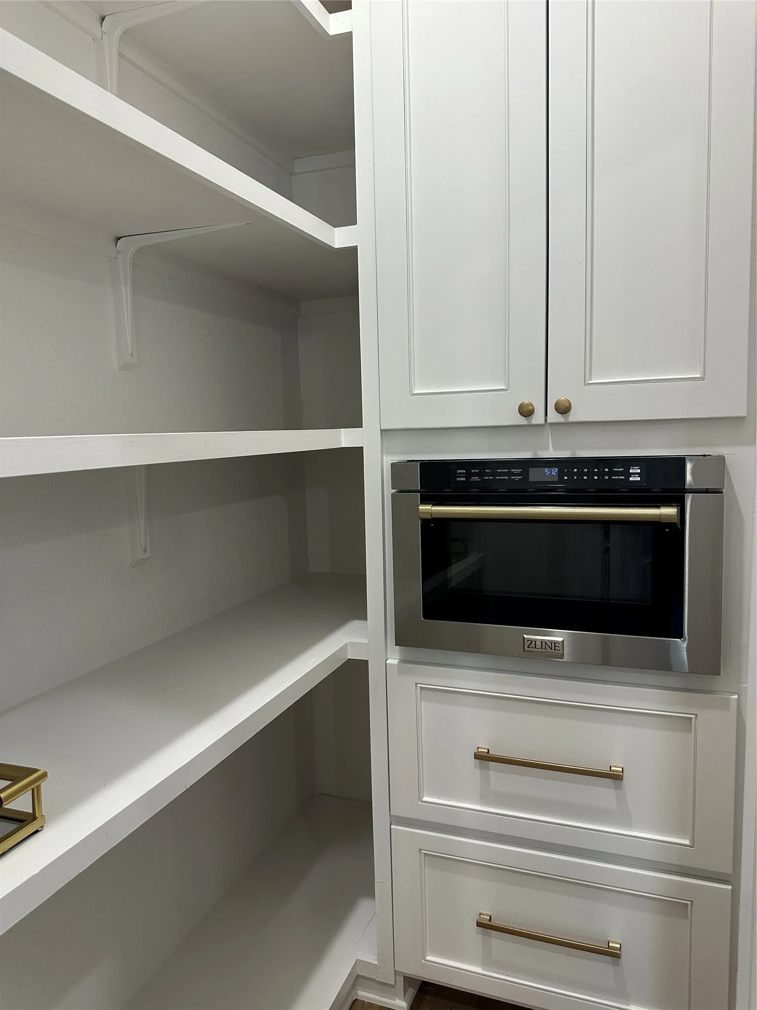 Built-in stainless steel microwave oven, white shaker-style cabinetry with gold-tone hardware, and extensive white shelving