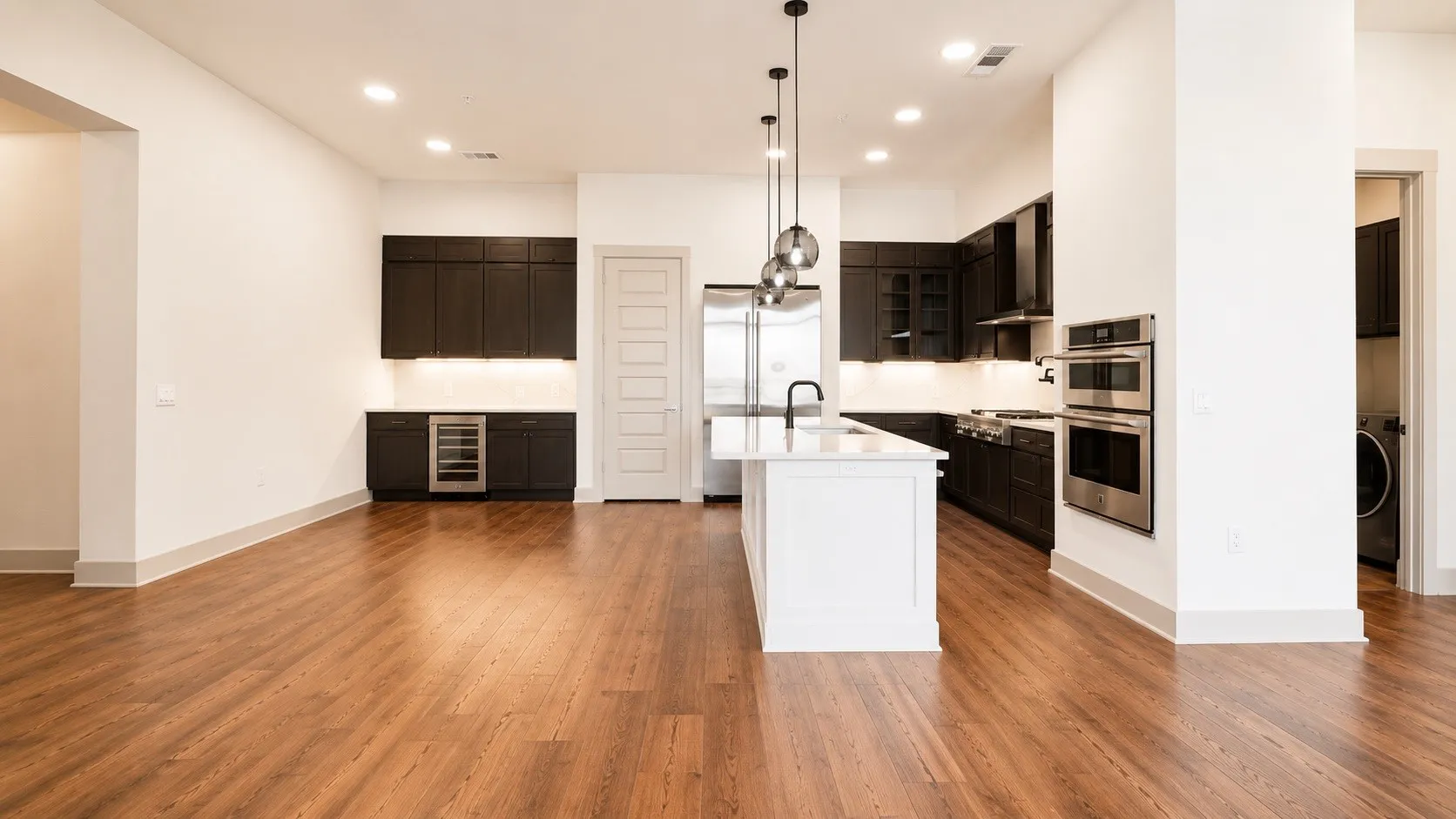 Open-concept kitchen featuring wood-finish flooring, a central island with a white countertop, dark cabinetry, and stainless steel appliances, including double wall ovens