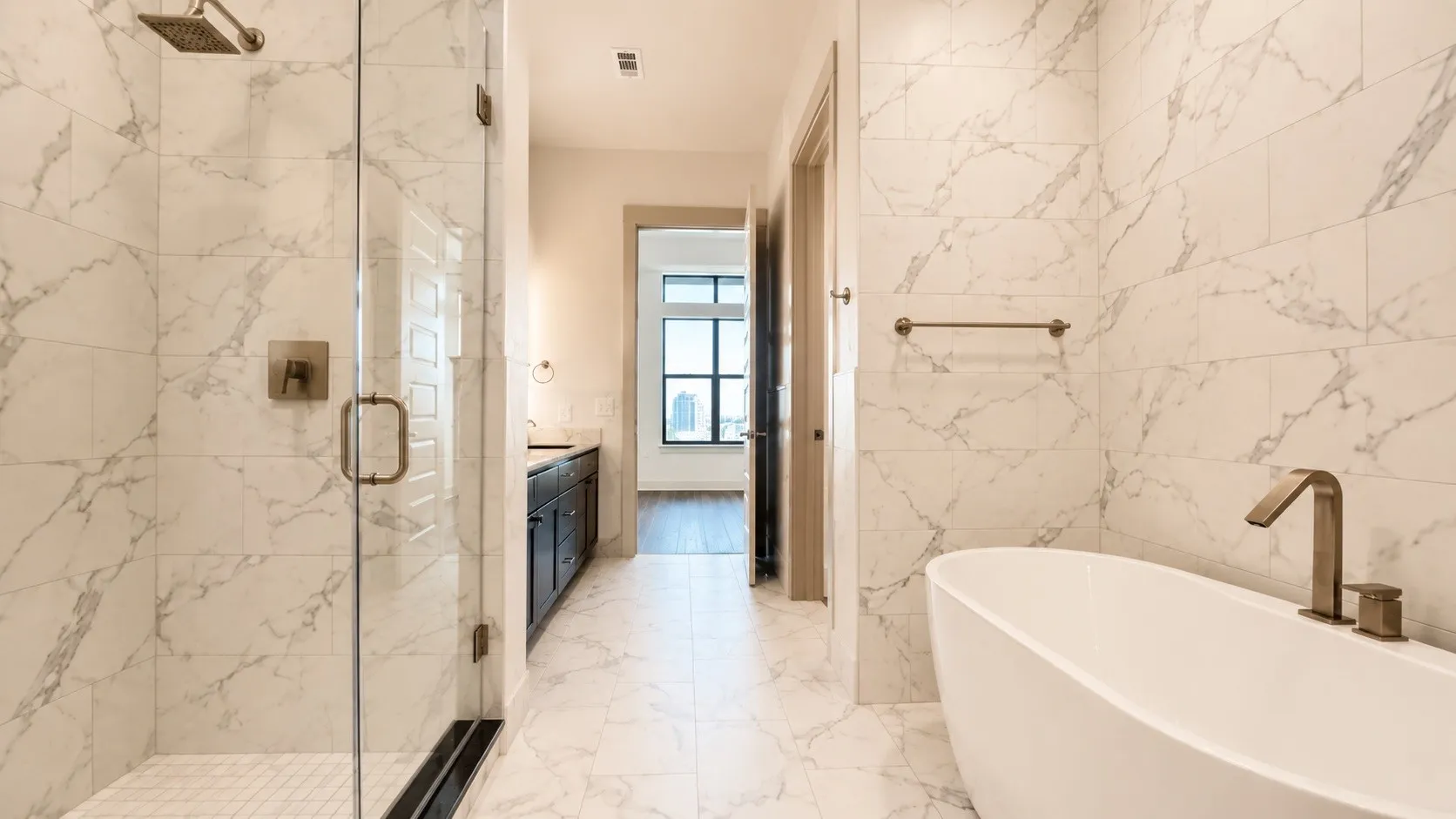 Spacious bathroom featuring marble-look tile flooring and walls, a freestanding soaking tub with bronze-finish fixtures, and a frameless glass shower enclosure with a rain showerhead