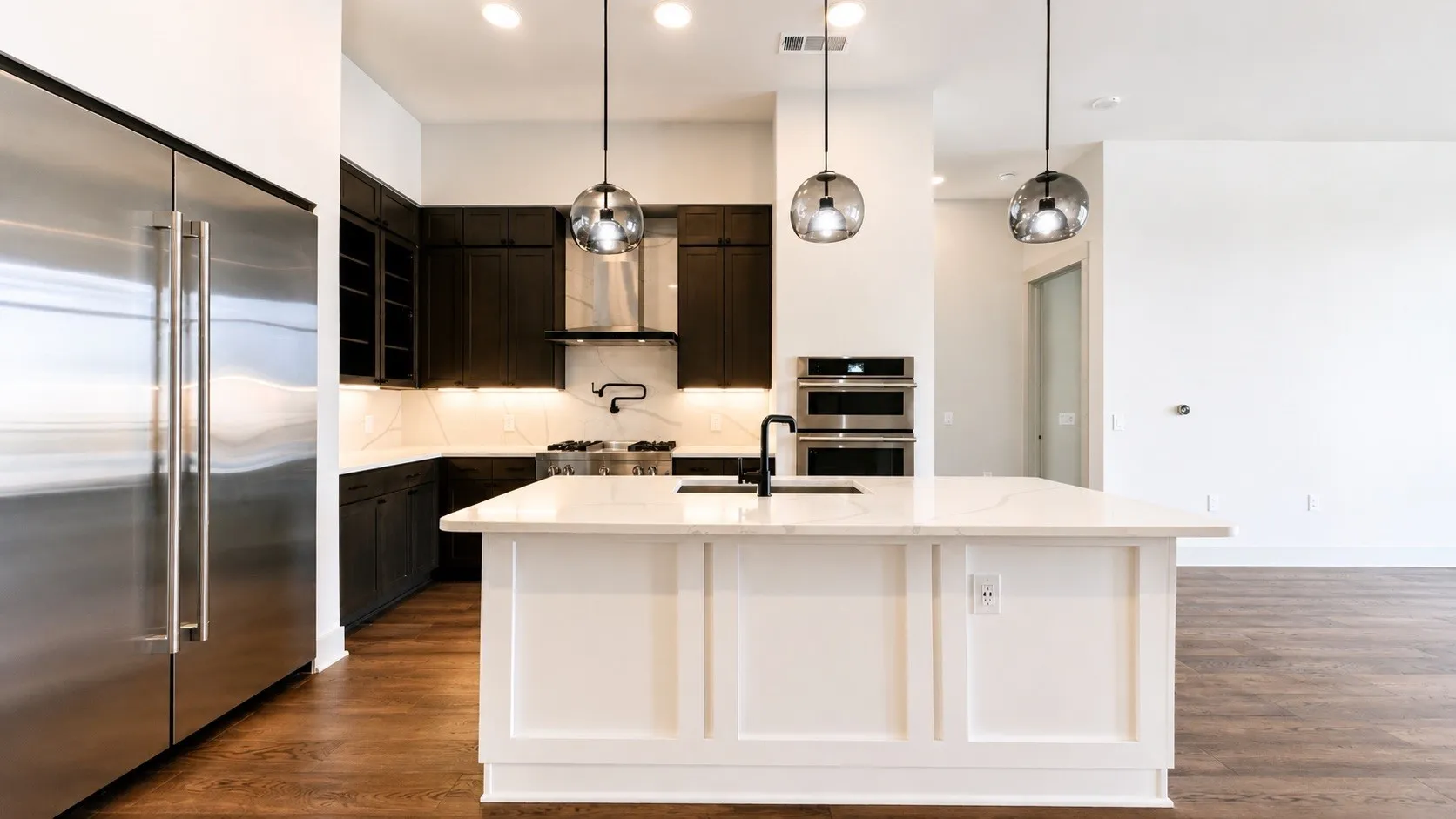 Modern kitchen featuring a large white island with a matte black faucet, dark wood cabinetry, stainless steel appliances, and wood-finish flooring