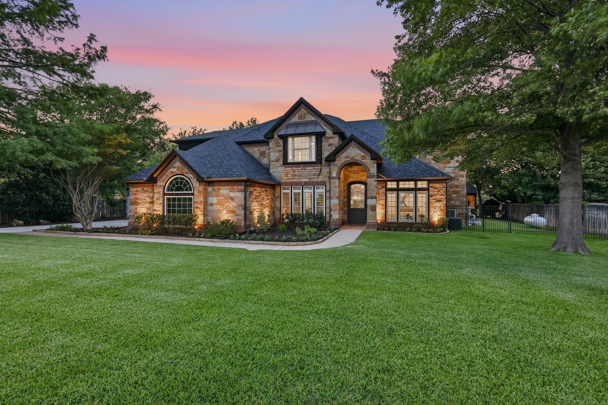 210 Canyon Oaks, Argyle, TX 76226 - Front of Home at Twilight - Stone and brick exterior residence featuring a prominent arched entry, black-framed windows, and a dark shingle roof