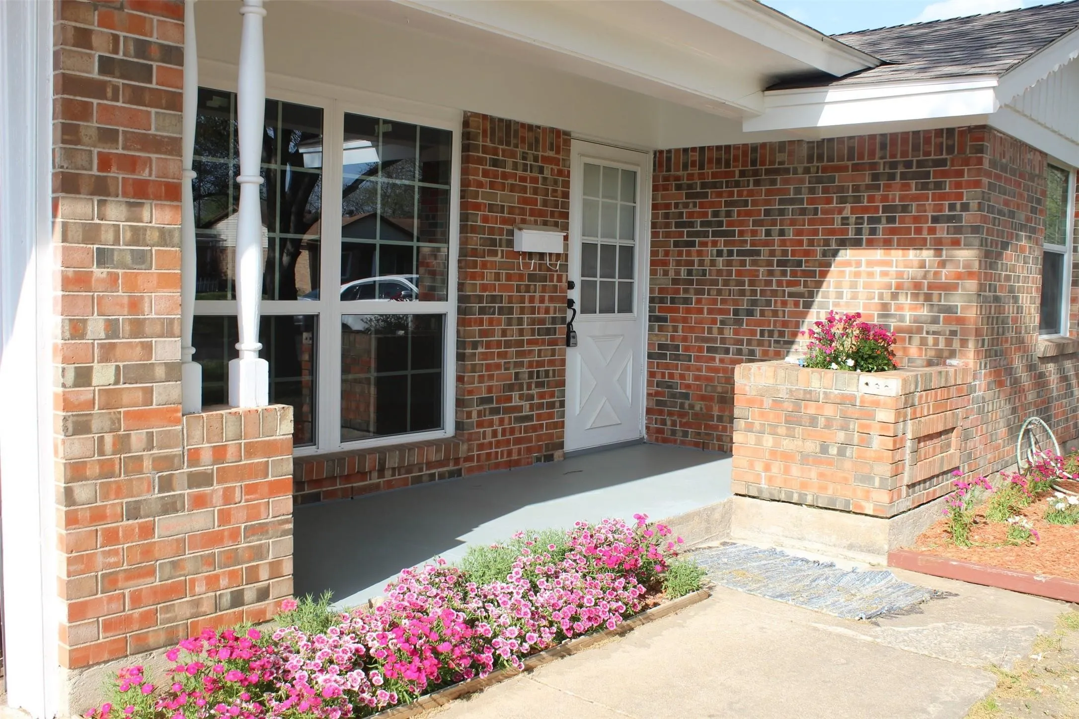 Brick exterior featuring a covered porch with white columns, a gridded picture window, a white paneled entry door, and a brick planter box with colorful landscaping