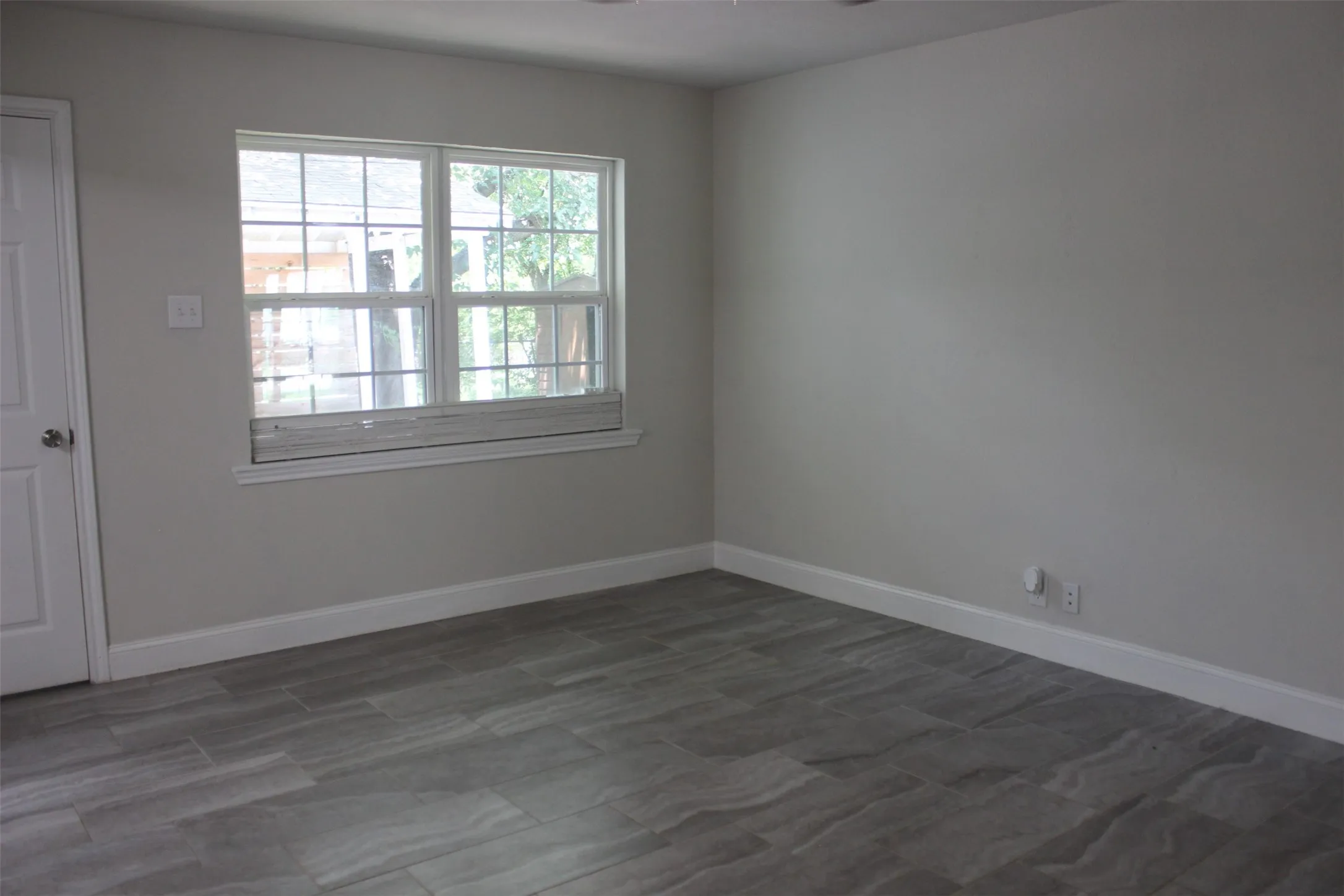 Spacious room featuring wood-finish tile flooring, a large window with white trim, and light gray painted walls. It also has a door to the back yard.