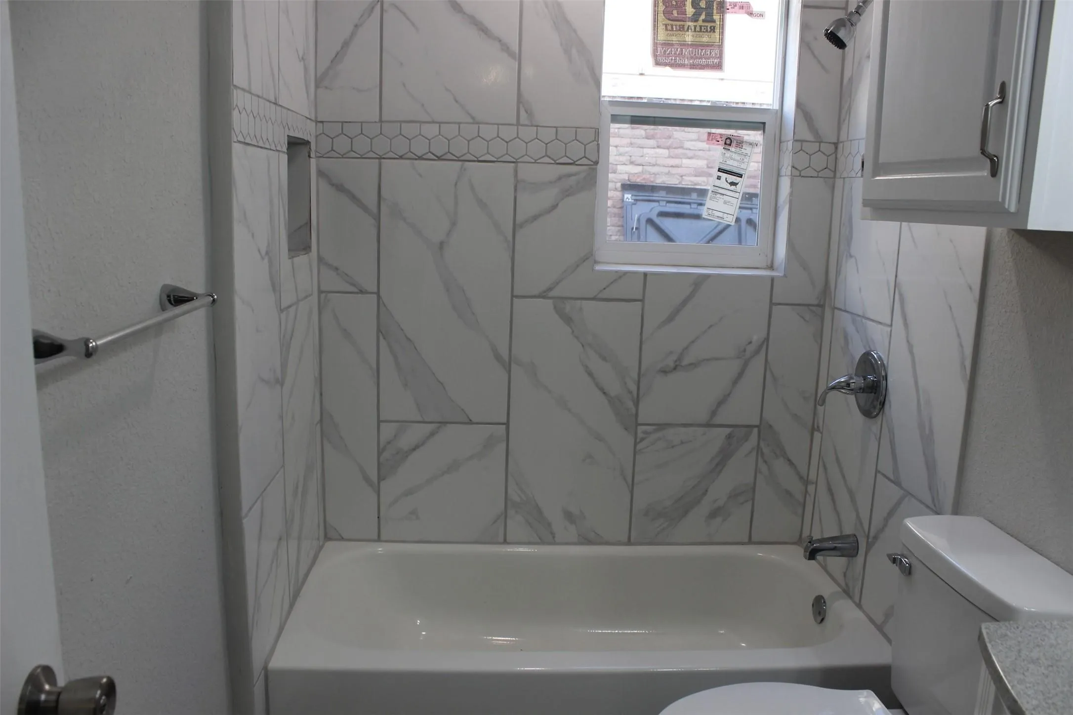 Bathroom featuring a white porcelain bathtub with a shower, white-tiled walls with a marble-effect pattern, a hexagonal mosaic tile accent band, and a recessed shower niche