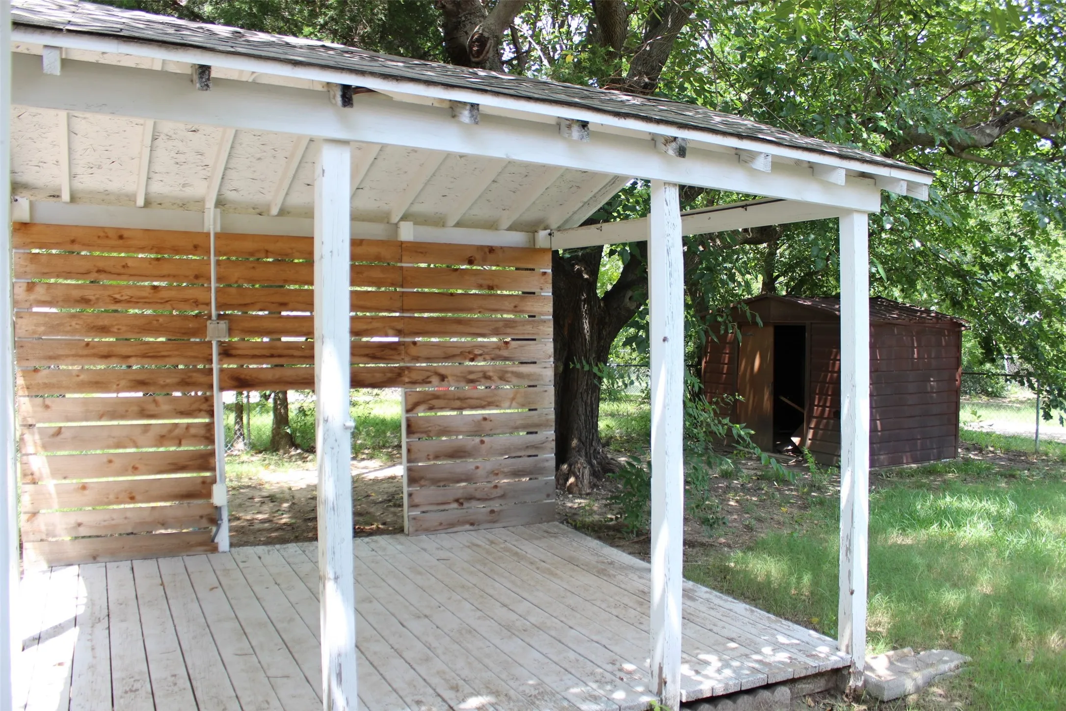 Covered outdoor structure featuring wood-plank deck, white support posts, and a partial wood-slat wall. You can grill or just relax in the backyard