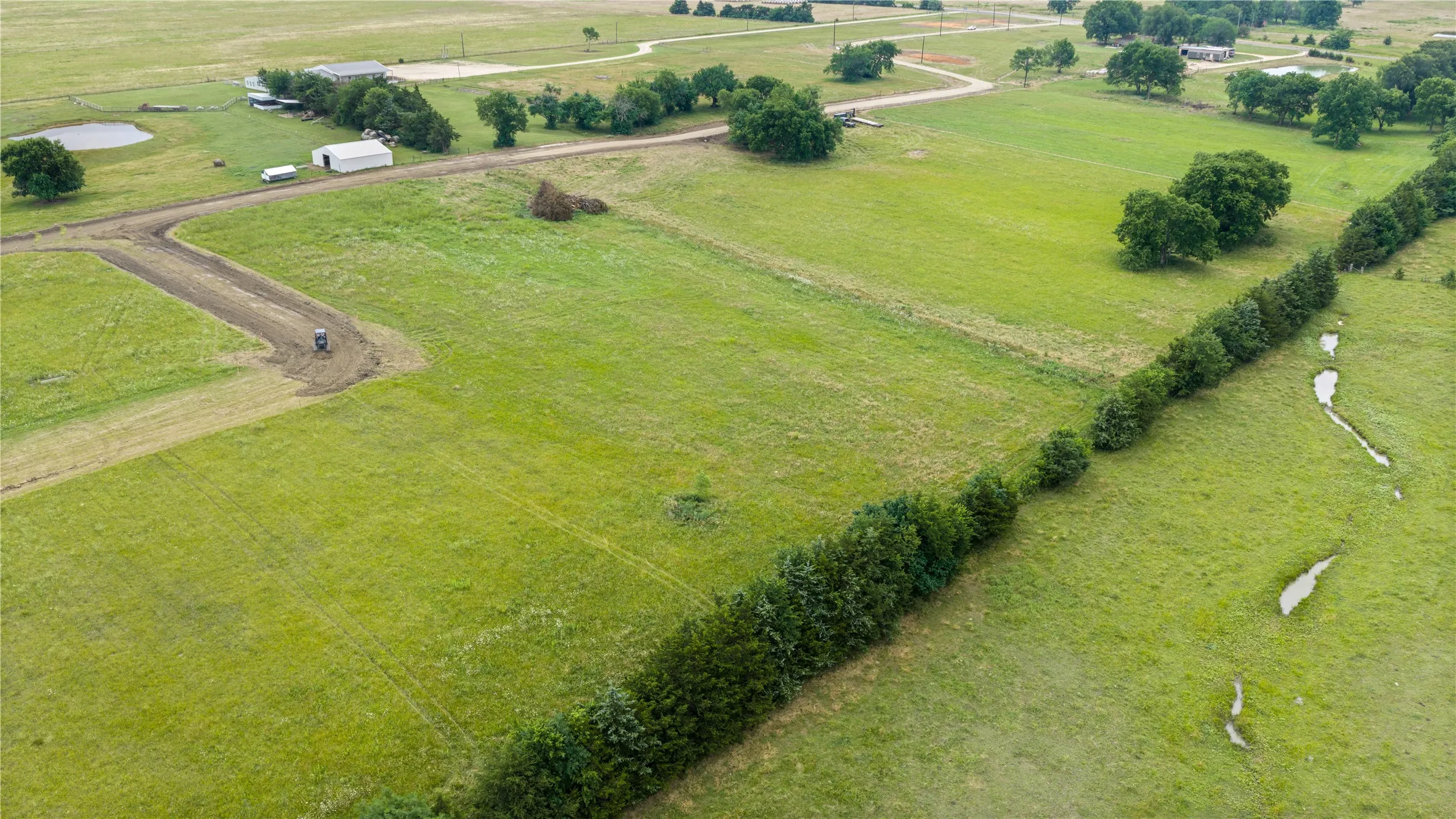Expansive green acreage featuring a newly graded dirt road, mature treeline, and a visible pond