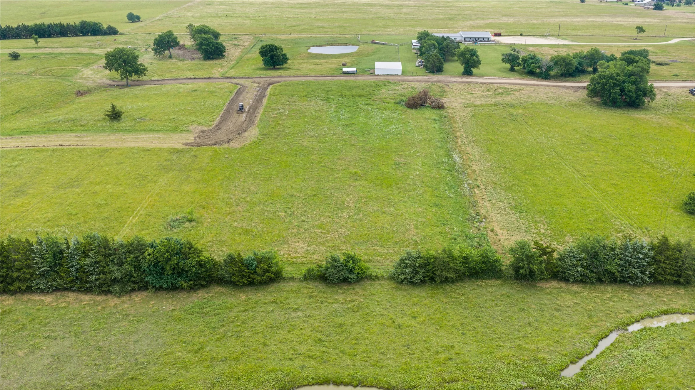Expansive green pastureland featuring a partial treeline, a small pond, and an unpaved access road