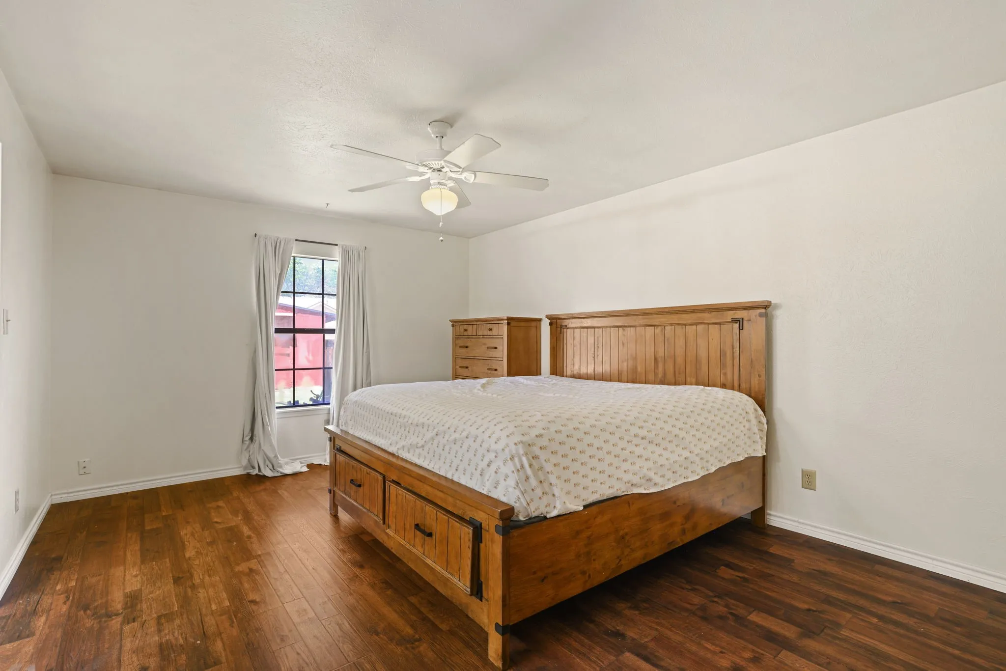 Spacious room featuring wood-finish flooring, a window with natural light, a ceiling fan with integrated lighting, and white baseboards