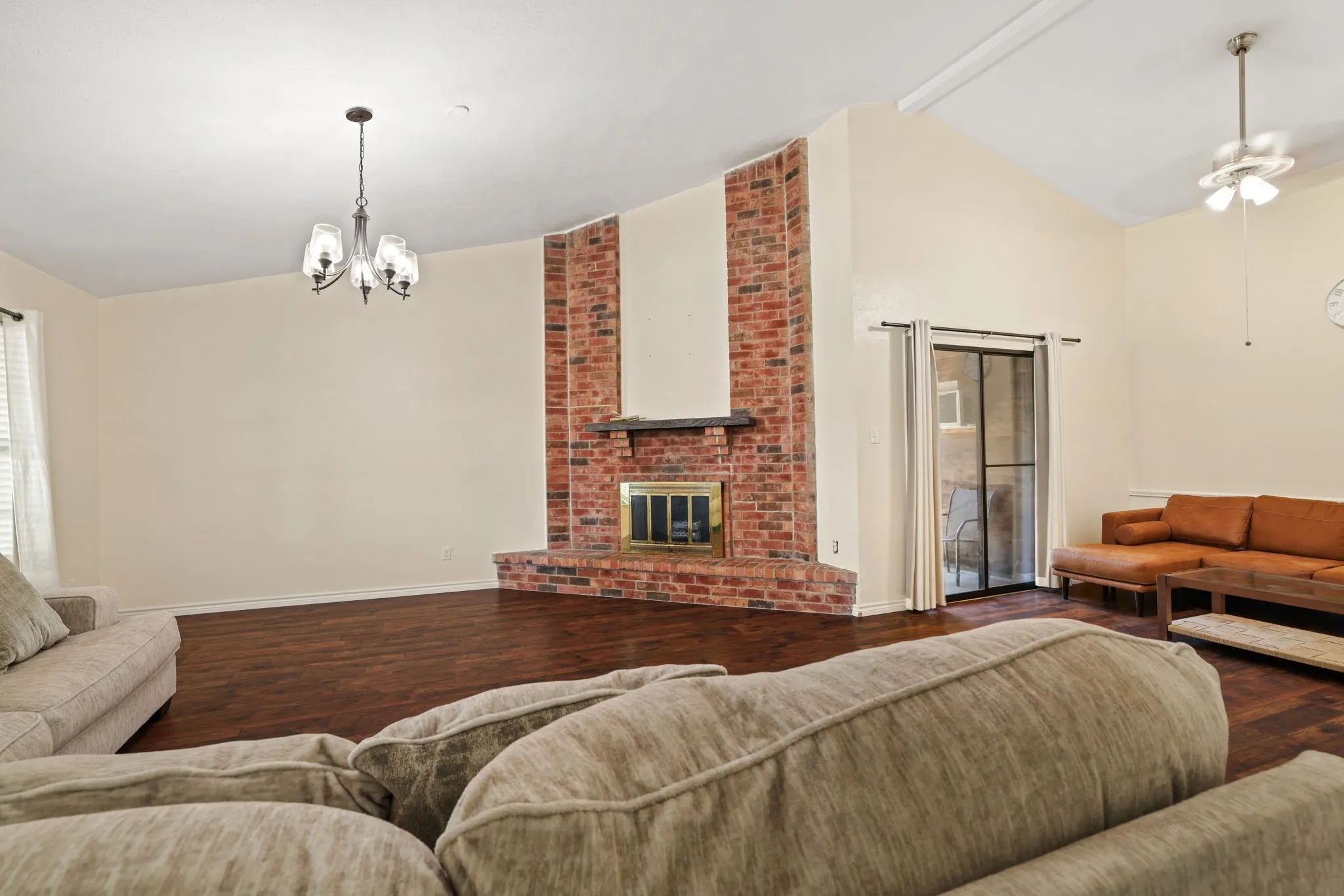 Spacious living area featuring a prominent brick fireplace with a dark mantel, wood-finish flooring, and a vaulted ceiling