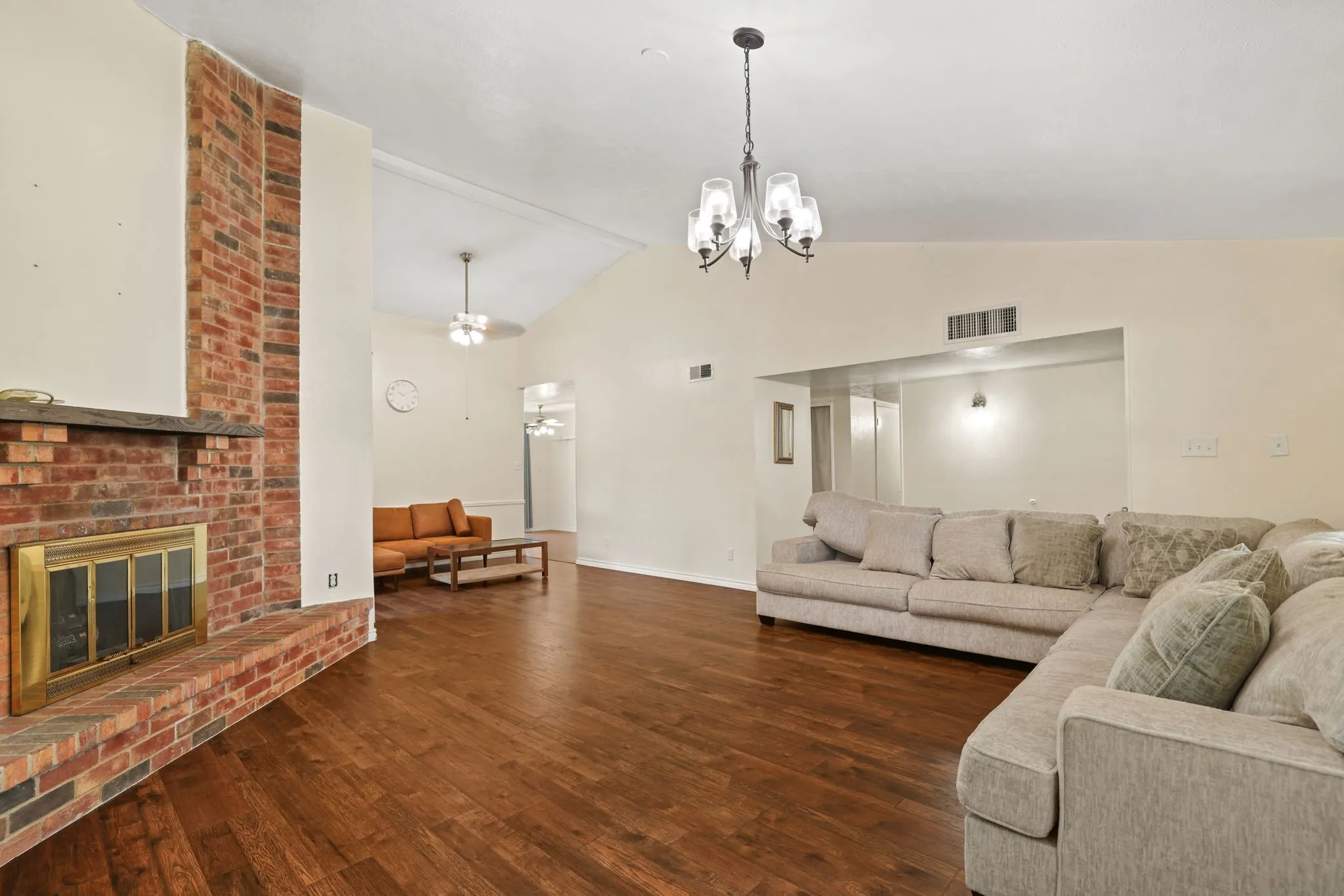 Spacious living area featuring a prominent brick fireplace with a brass firebox, wood-finish flooring, and a vaulted ceiling with a ceiling fan