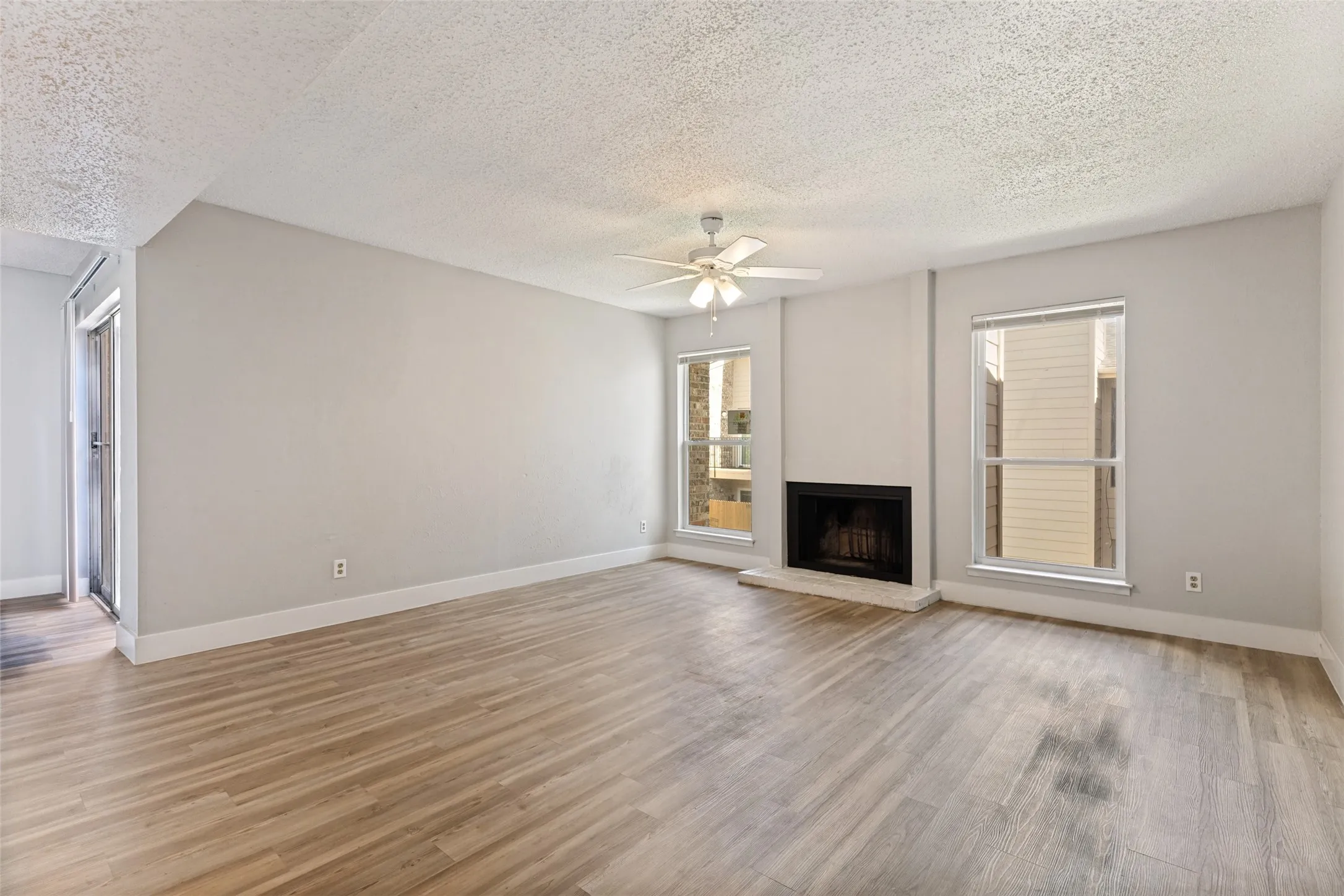Spacious living area featuring wood-finish flooring, a built-in fireplace with a stone hearth, two large windows, a ceiling fan with integrated lighting, and neutral wall tones