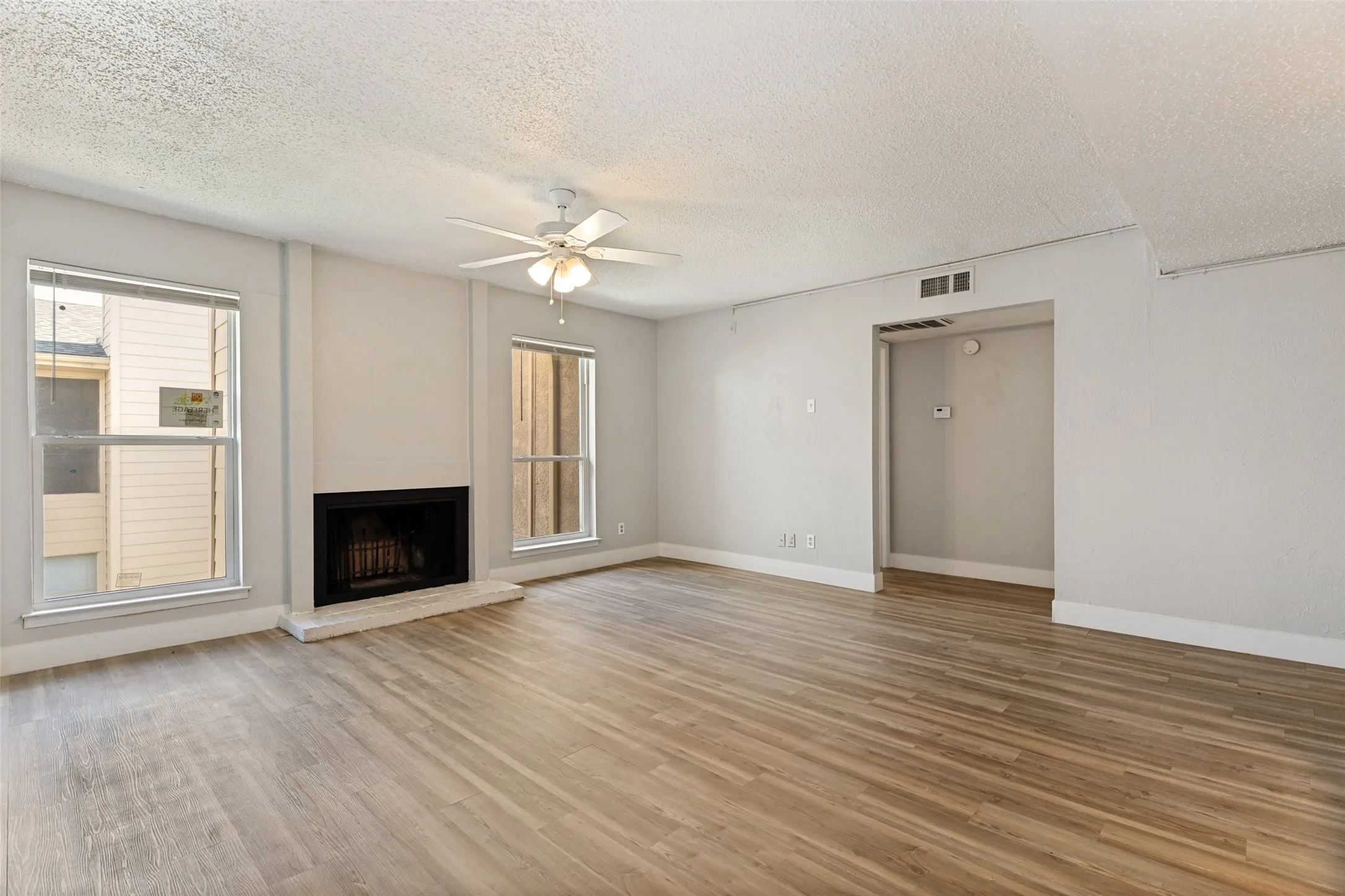 Spacious living area featuring wood-finish flooring, a fireplace with a light-toned hearth, and two windows providing natural illumination