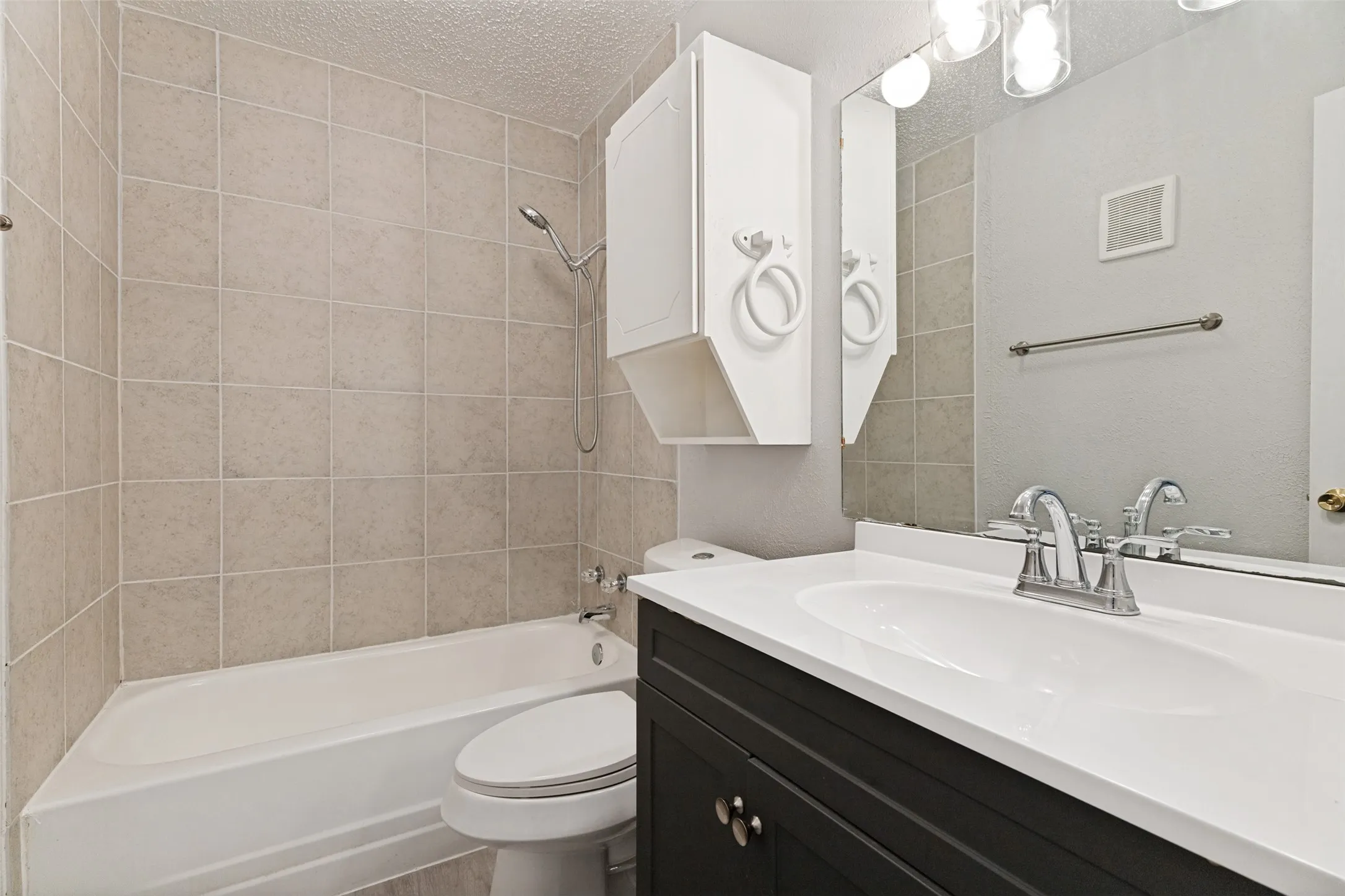 Full bathroom featuring a white tub, light-toned tile surround, a built-in white storage cabinet, and a vanity with a dark cabinet base, white countertop, and integrated sink