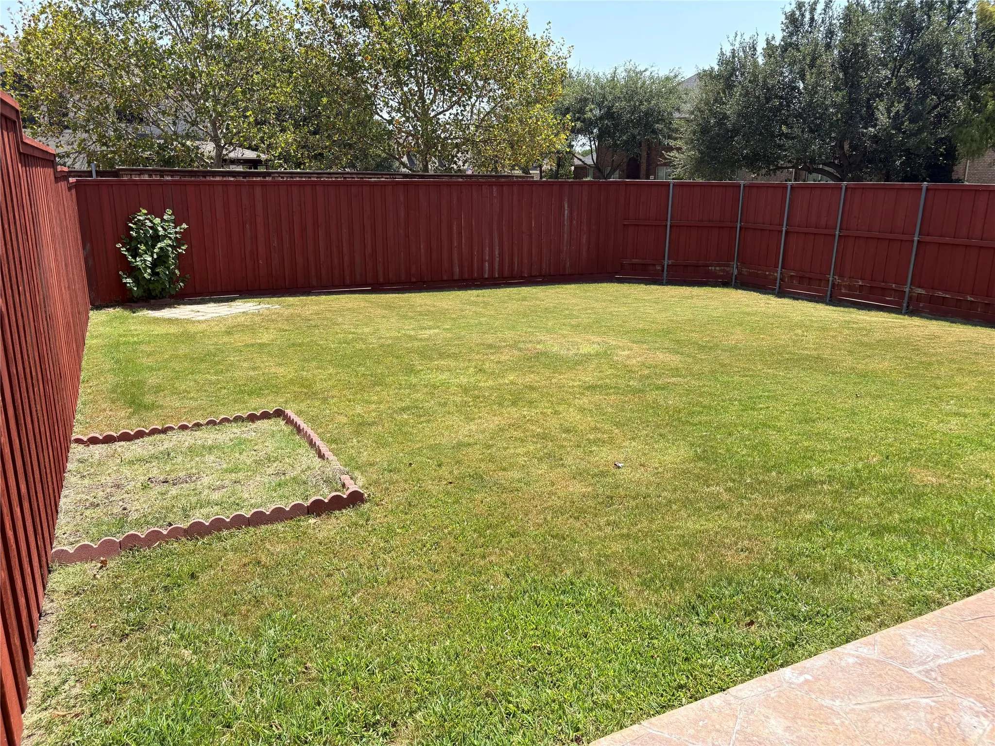 Spacious backyard featuring a large grass lawn, perimeter wood fencing, decorative garden edging, and a mature shrub
