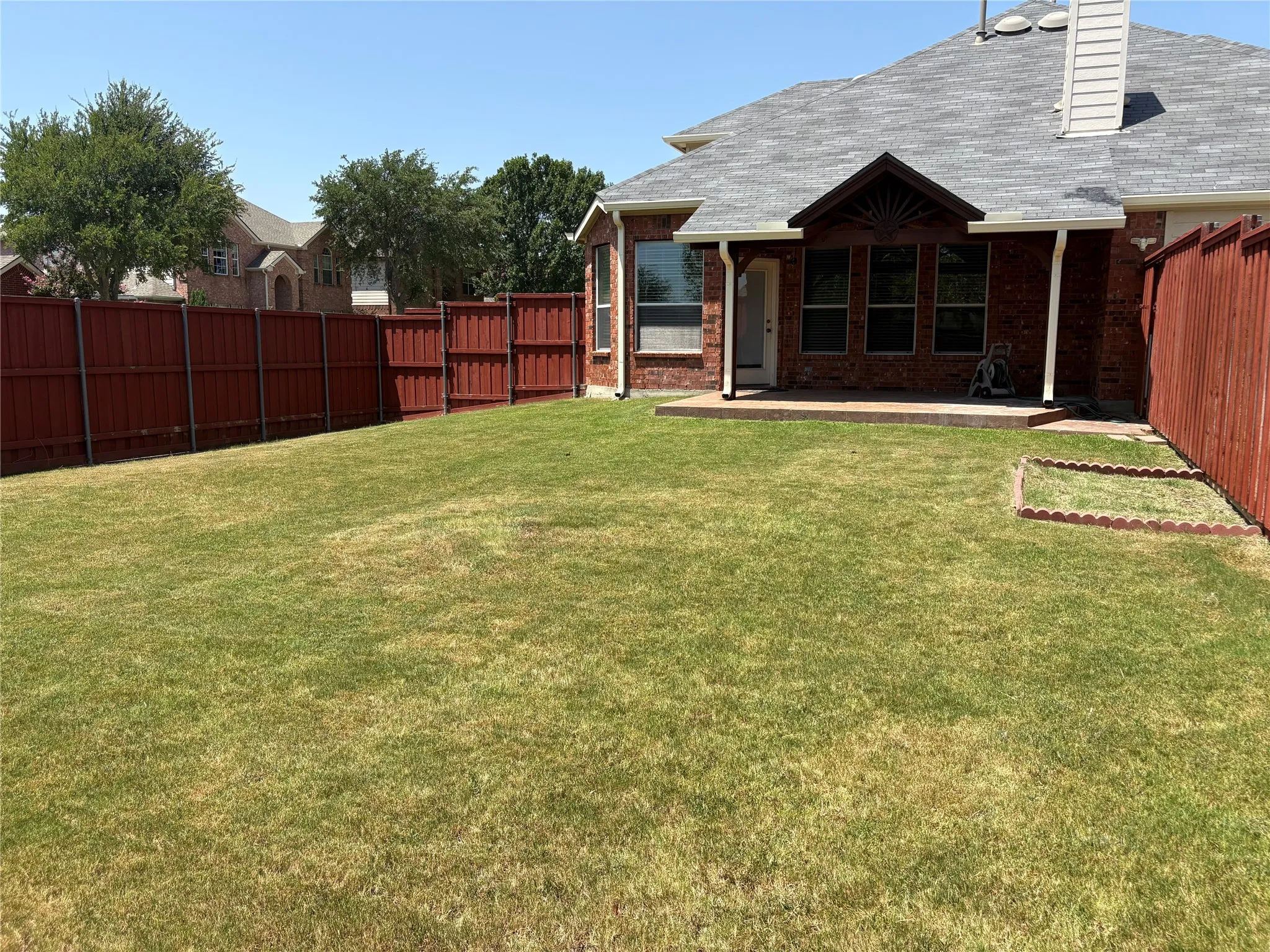 Expansive backyard featuring a manicured lawn, full privacy fencing, a covered patio, brick exterior, and a gabled roofline