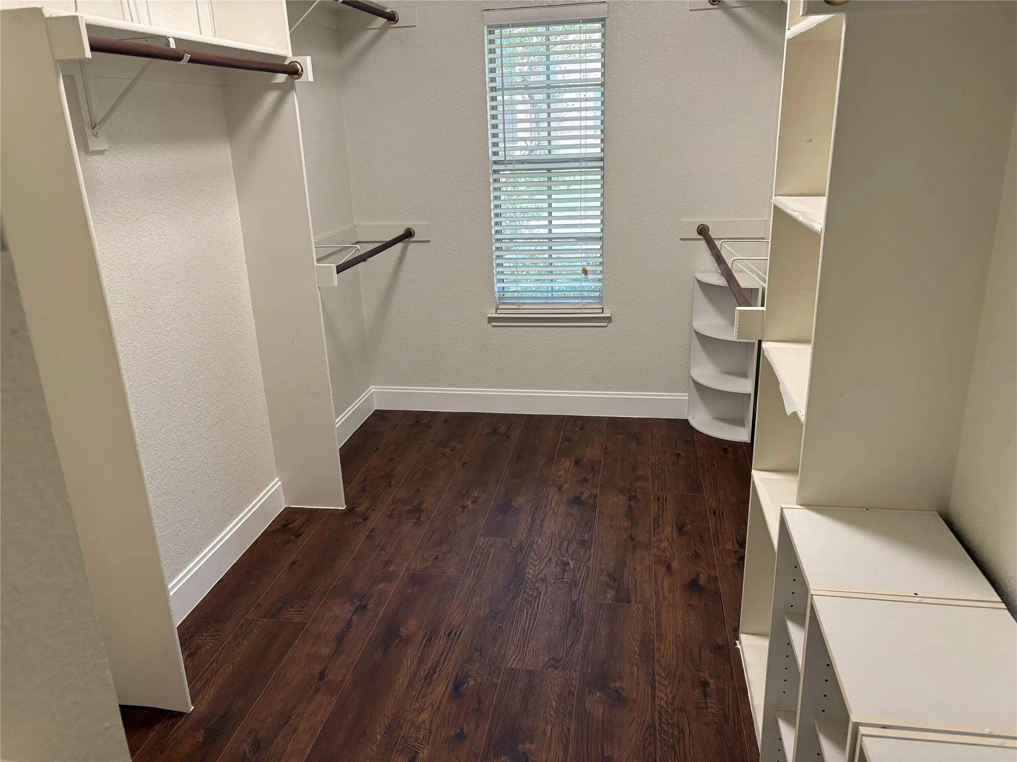 Spacious walk-in closet featuring extensive built-in shelving, hanging rods, wood-finish flooring, a window with blinds, and white baseboards