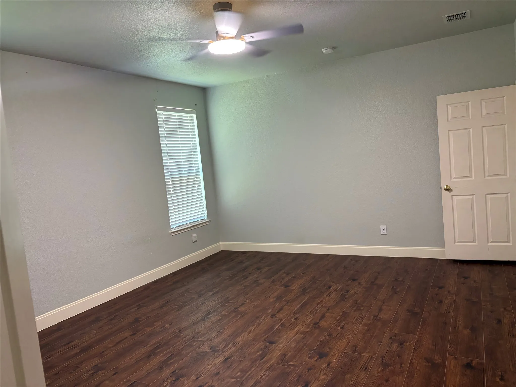 Spacious room featuring wood-finish flooring, a ceiling fan with integrated lighting, a single window with blinds, light gray wall paint, and white trim
