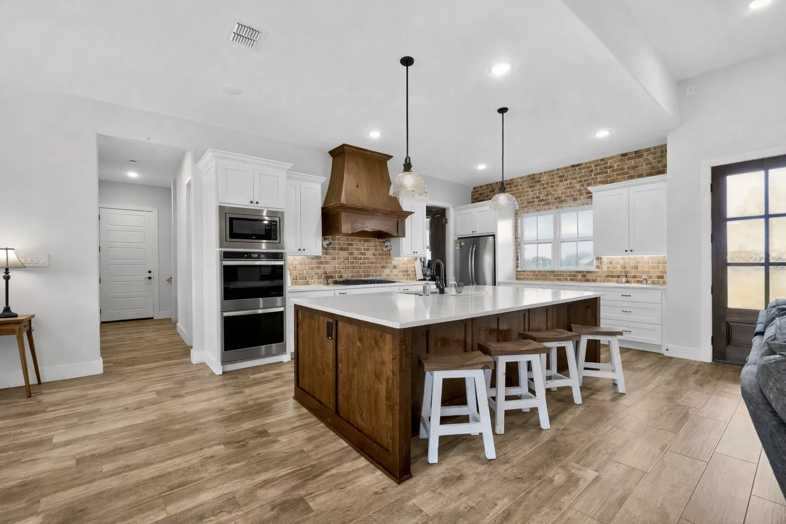 Gourmet kitchen with a large central island featuring a white countertop and wood-finish cabinetry, integrated stainless steel appliances, white upper and lower cabinetry, a brick-finish backsplash, and wood-finish flooring