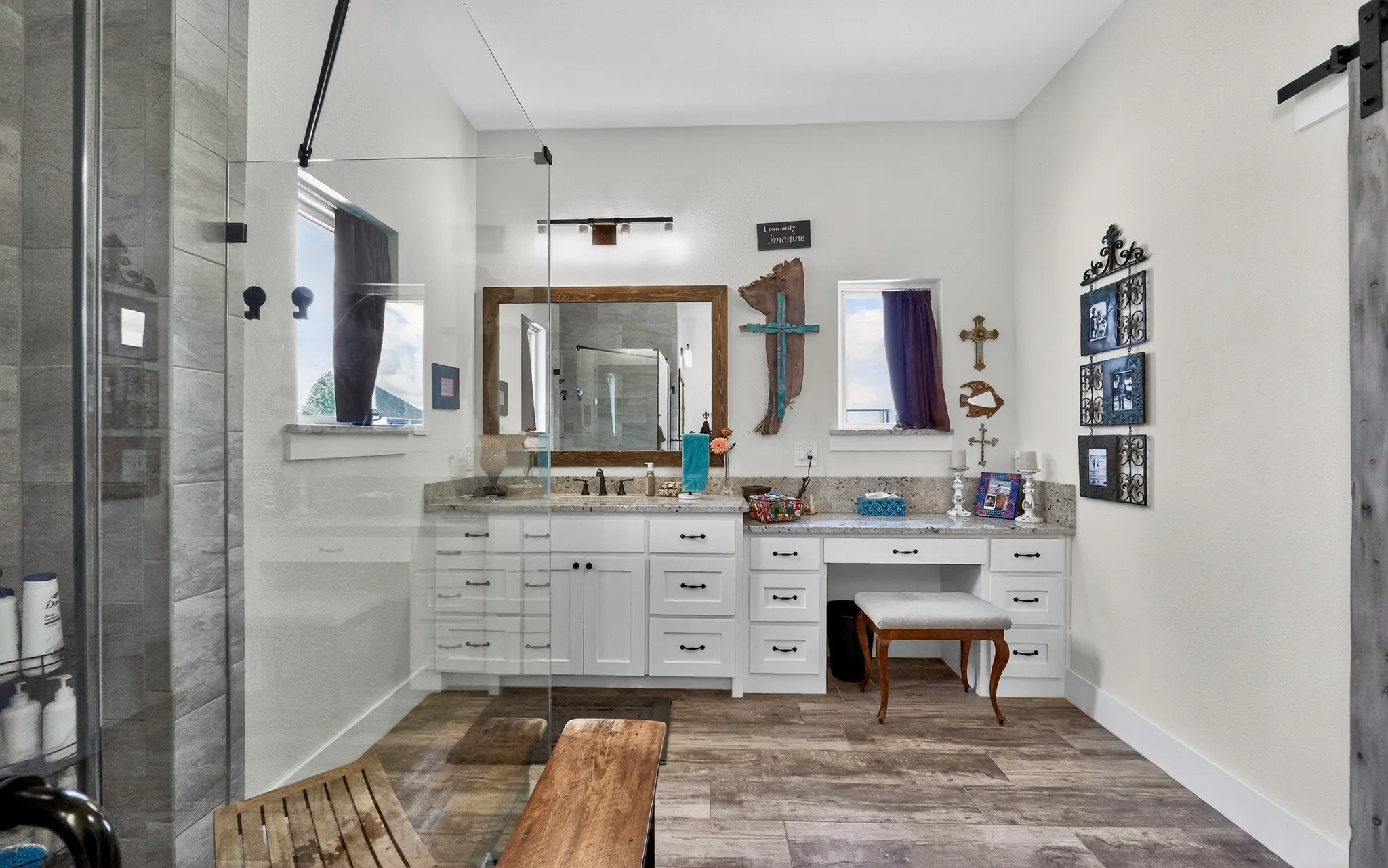 Spacious bathroom featuring a frameless glass shower enclosure with gray tile surround, wood-finish flooring, a double vanity with white cabinetry and a light-toned countertop, and an integrated makeup vanity