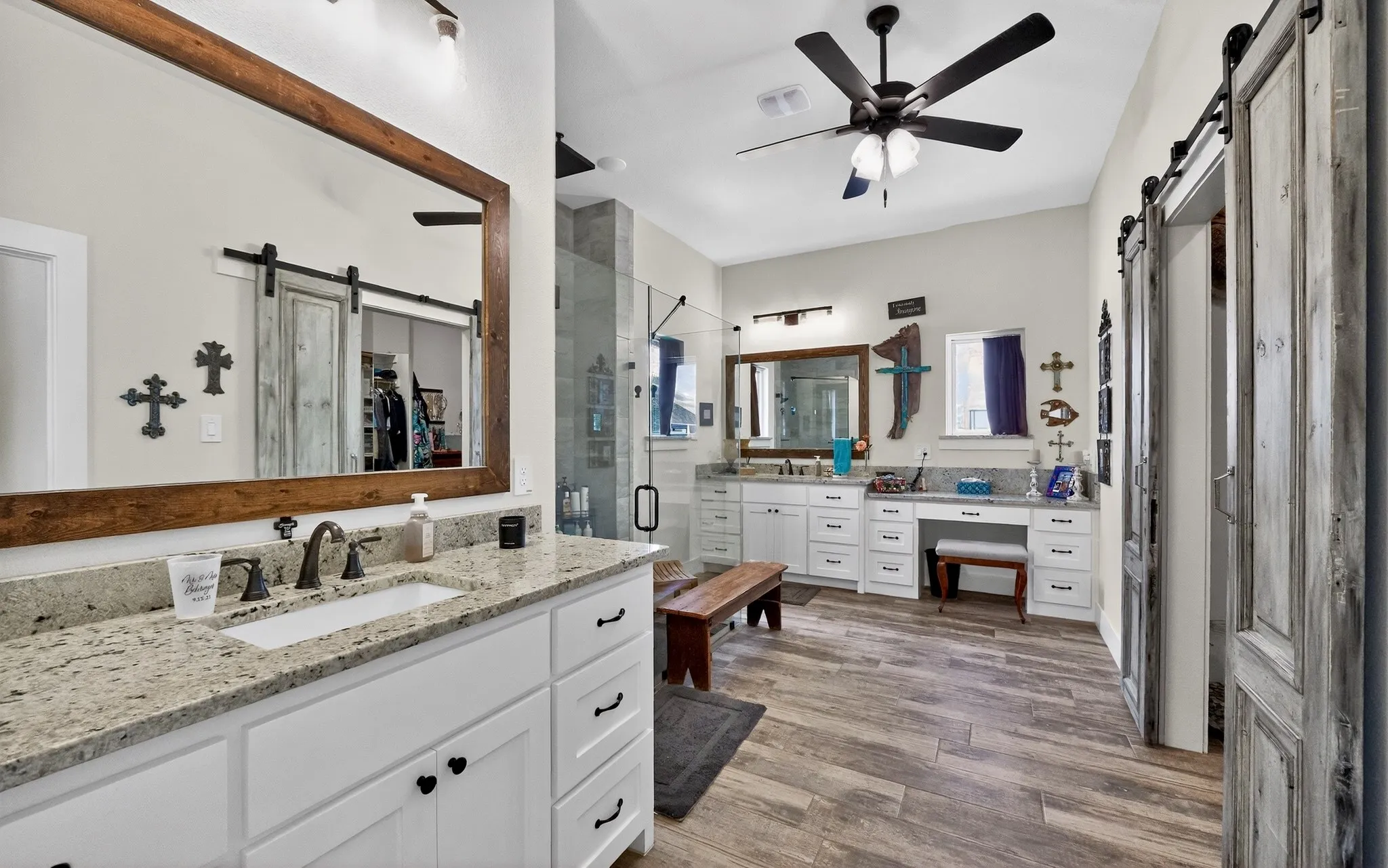 Spacious bathroom featuring wood-finish flooring, dual vanity areas with granite countertops, a large walk-in shower with glass enclosure, and sliding barn doors