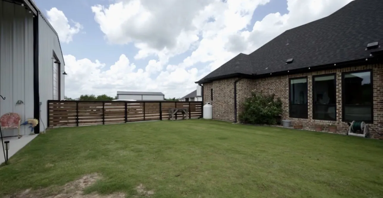 Expansive green lawn surrounds a brick exterior home with dark window frames and a shingled roof, complemented by a horizontal slat fence and an adjacent metal-sided outbuilding