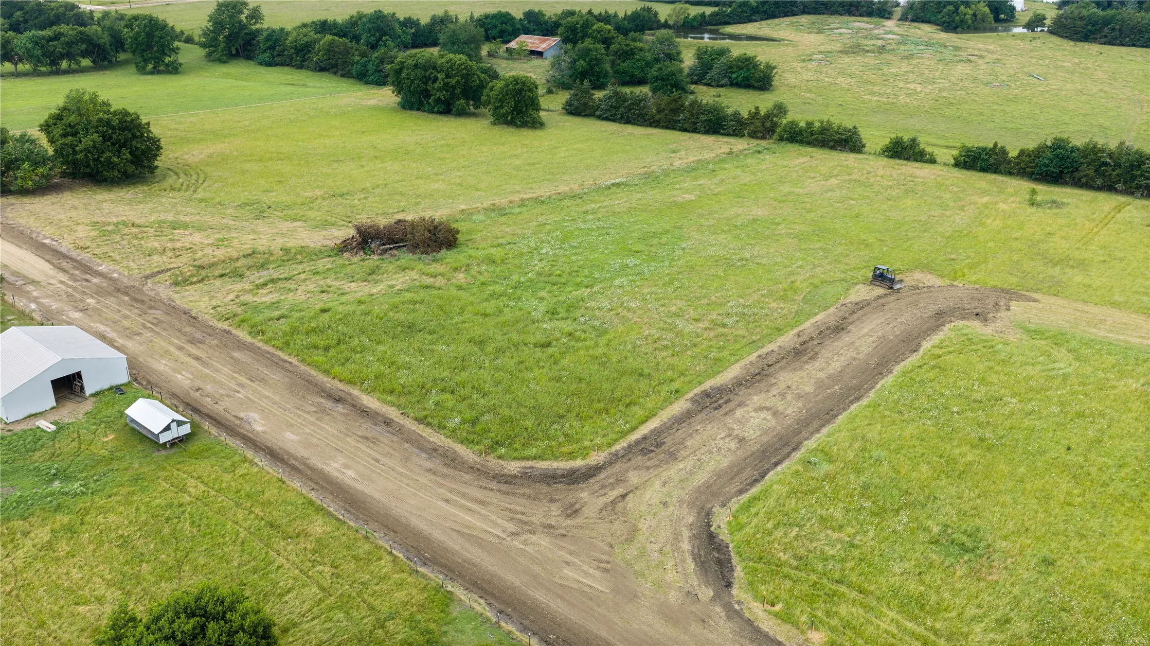 Expansive green fields featuring a metal barn structure, a cleared dirt path, and scattered mature trees