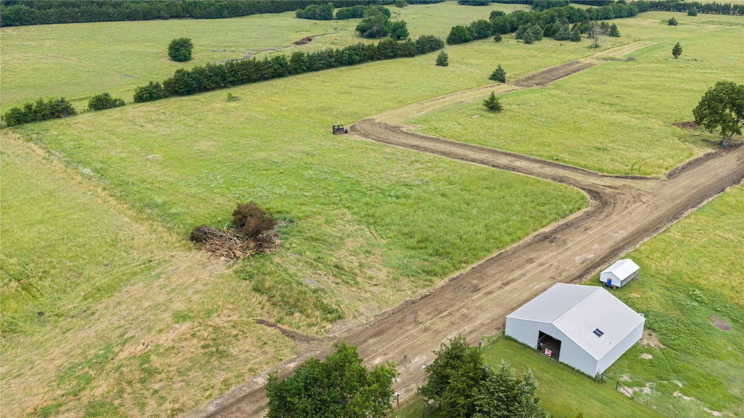 Expansive agricultural land featuring a large metal barn, multiple dirt paths, verdant green fields, and mature trees