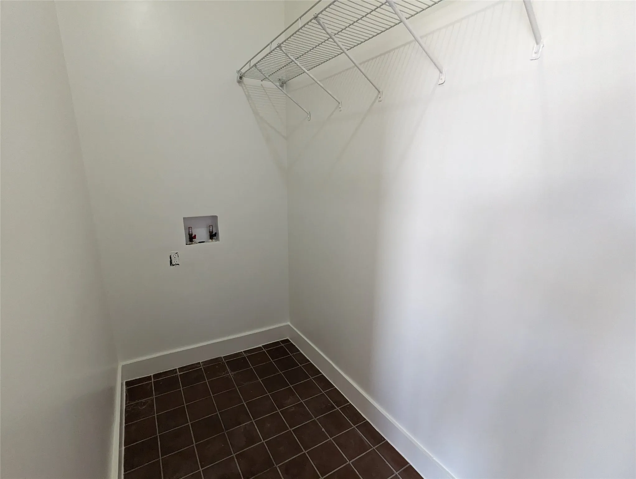 Utility area featuring brown square floor tiles, white baseboards, a wall-mounted wire shelf, and visible plumbing connections