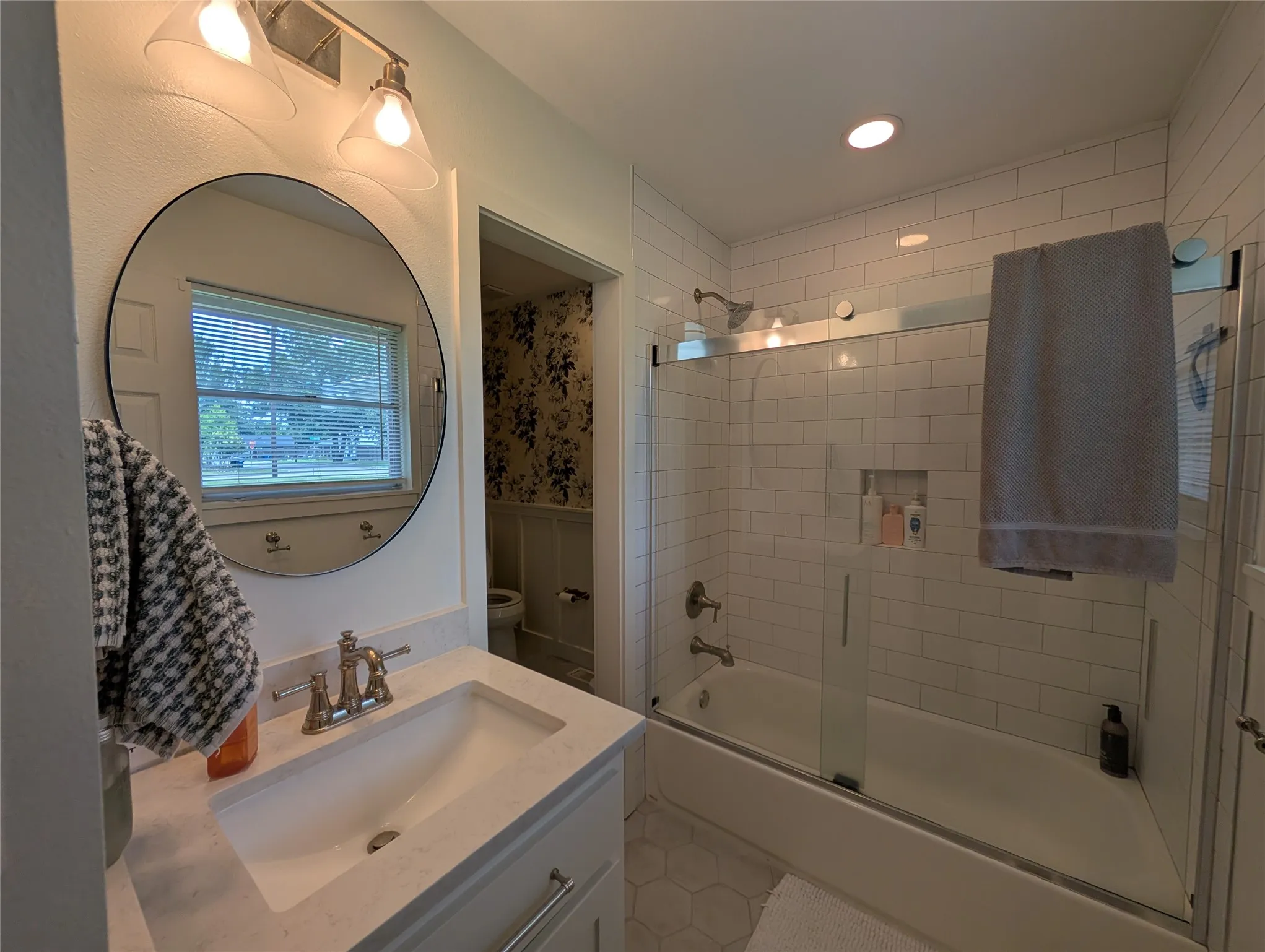 Bathroom featuring a vanity with an integrated sink and light-toned countertop, a large round mirror, and a shower-tub combination with a sliding glass door