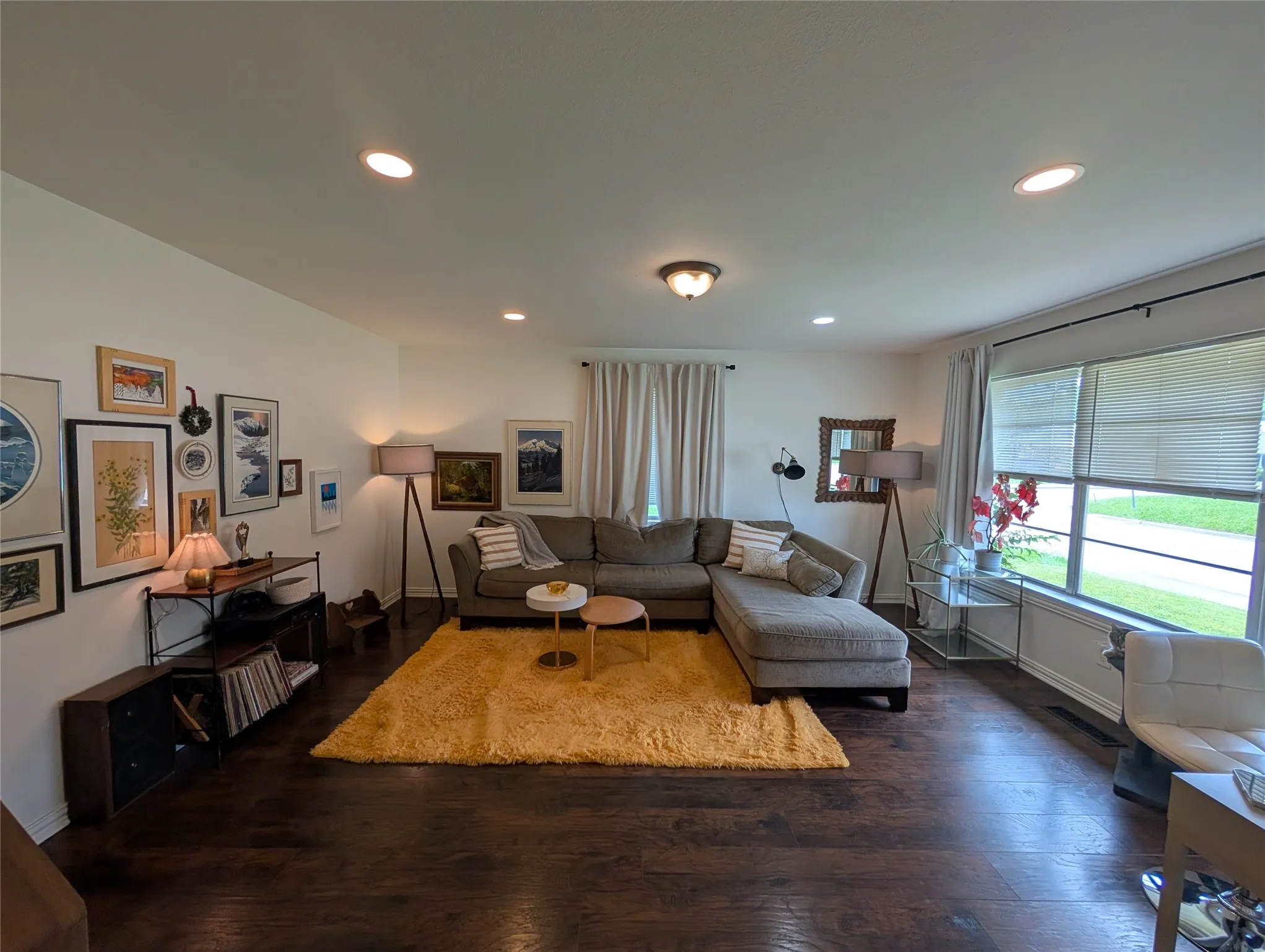 Spacious living area featuring rich wood-finish flooring, abundant recessed lighting, a large picture window with blinds and drapes, and an overhead flush-mount light fixture