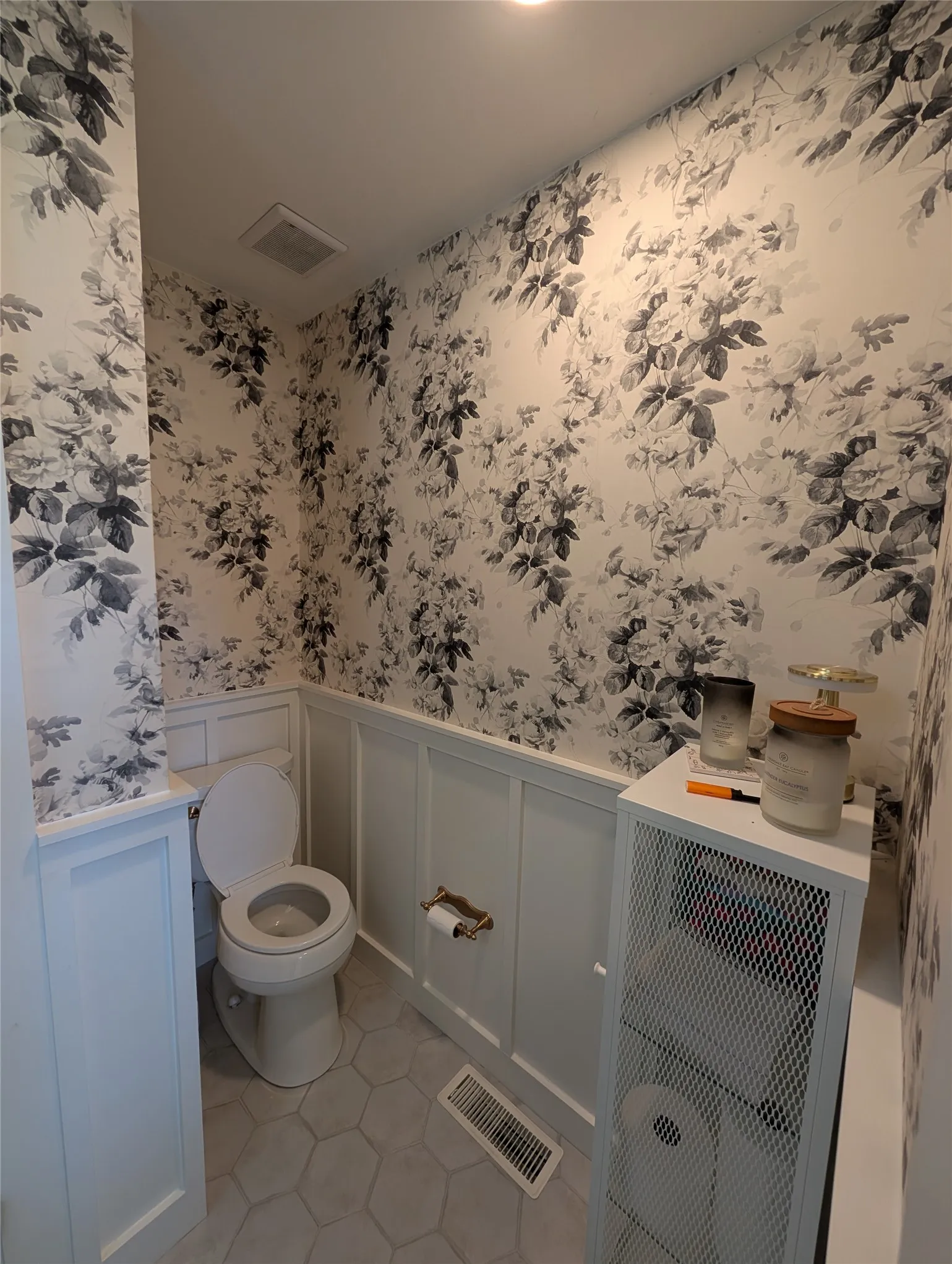 Powder room featuring botanical print wallpaper, white board-and-batten wainscoting, hexagon tile flooring, and a brass-tone toilet paper holder