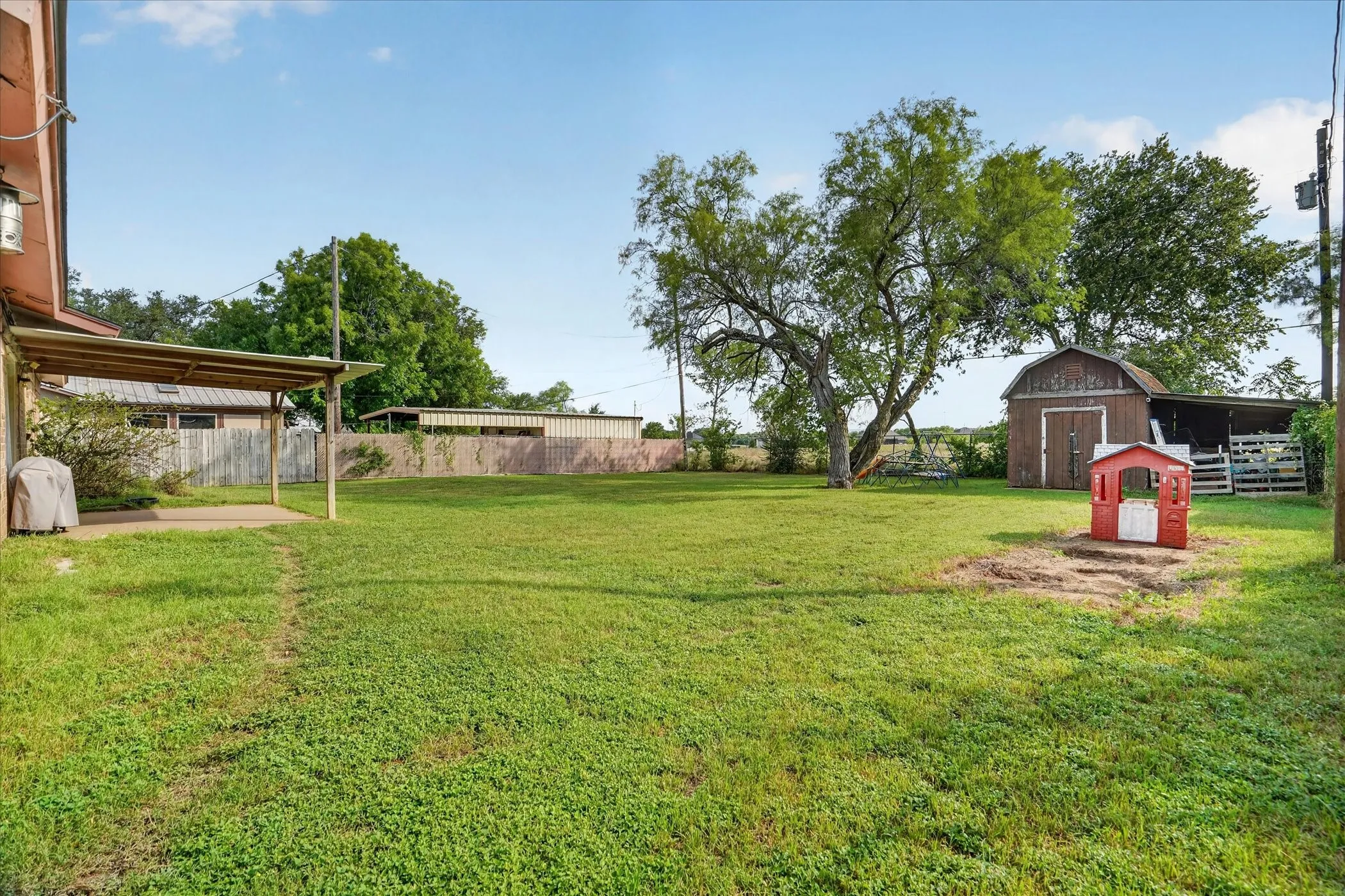 Expansive backyard featuring a covered concrete patio, mature trees, a wooden storage shed, and additional covered outdoor storage