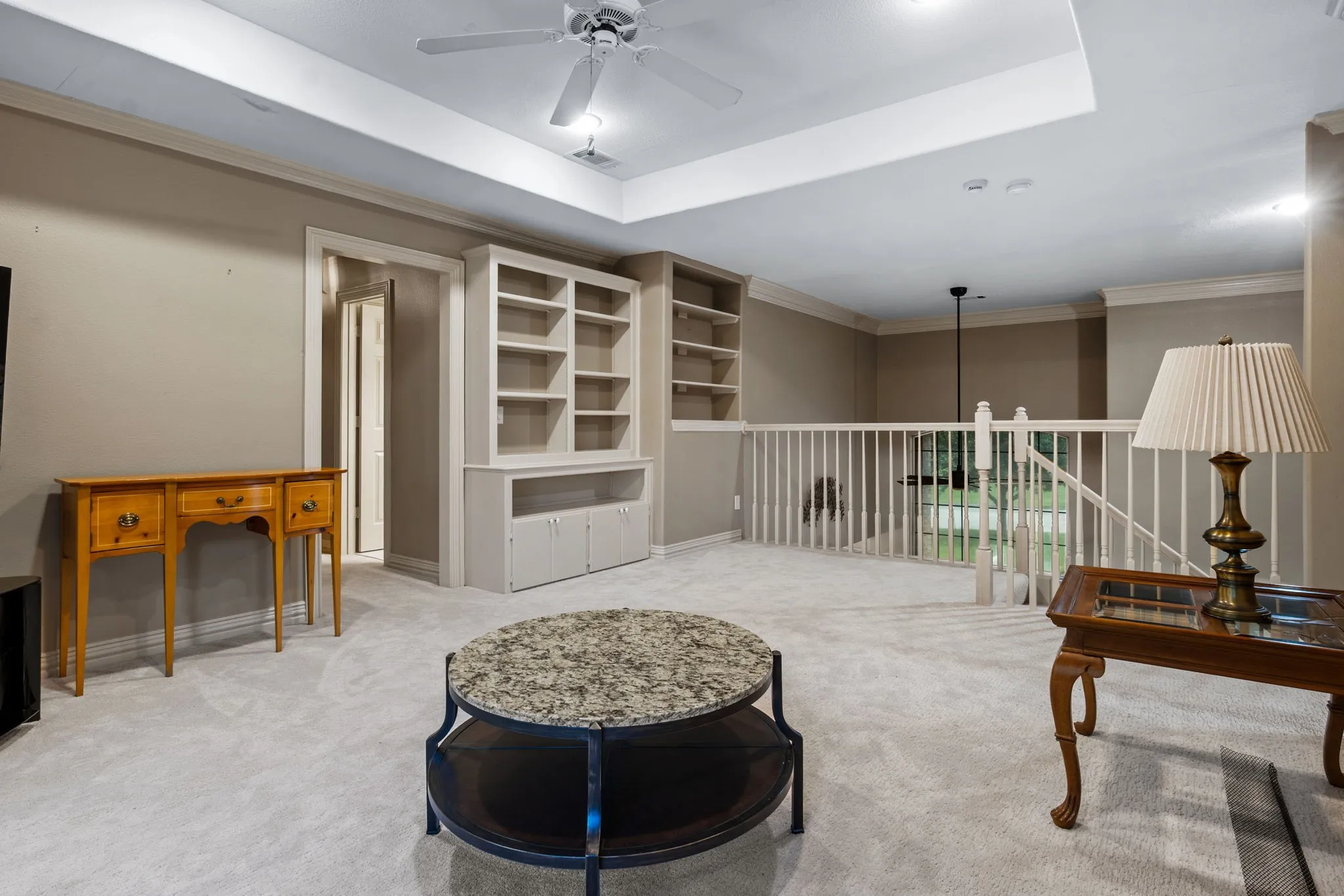 Spacious carpeted loft featuring a tray ceiling with a ceiling fan, built-in shelving, and a white railing overlooking the floor below