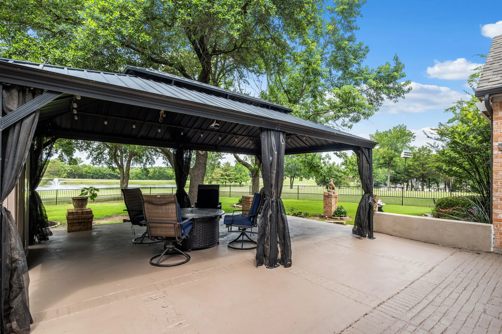 Expansive covered patio featuring a metal gazebo structure with black privacy curtains, overlooking a manicured lawn and a body of water with a fountain
